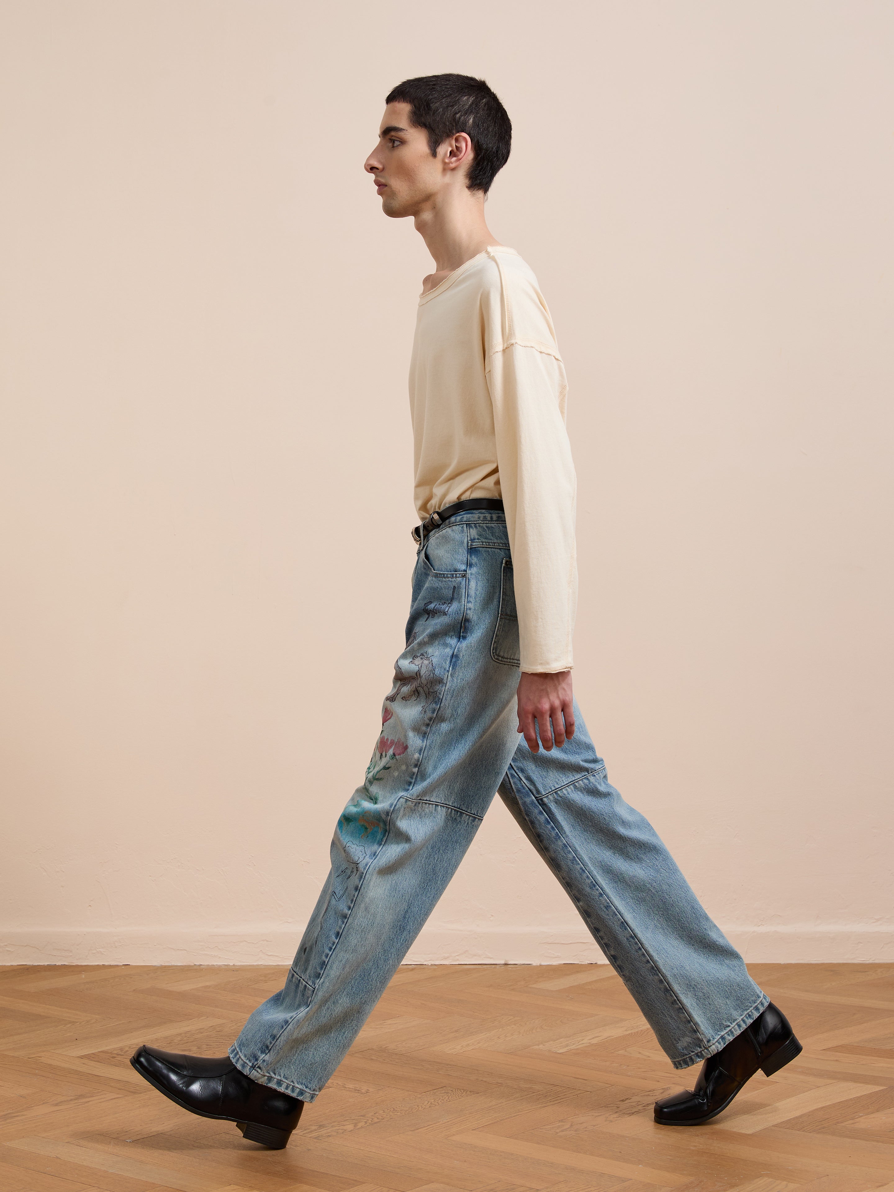 A person with short dark hair walks in profile on a wooden floor, wearing a cream long-sleeve shirt, black shoes, and FOUND’s Hand Art Jeans—blue denim jeans featuring paint marks.