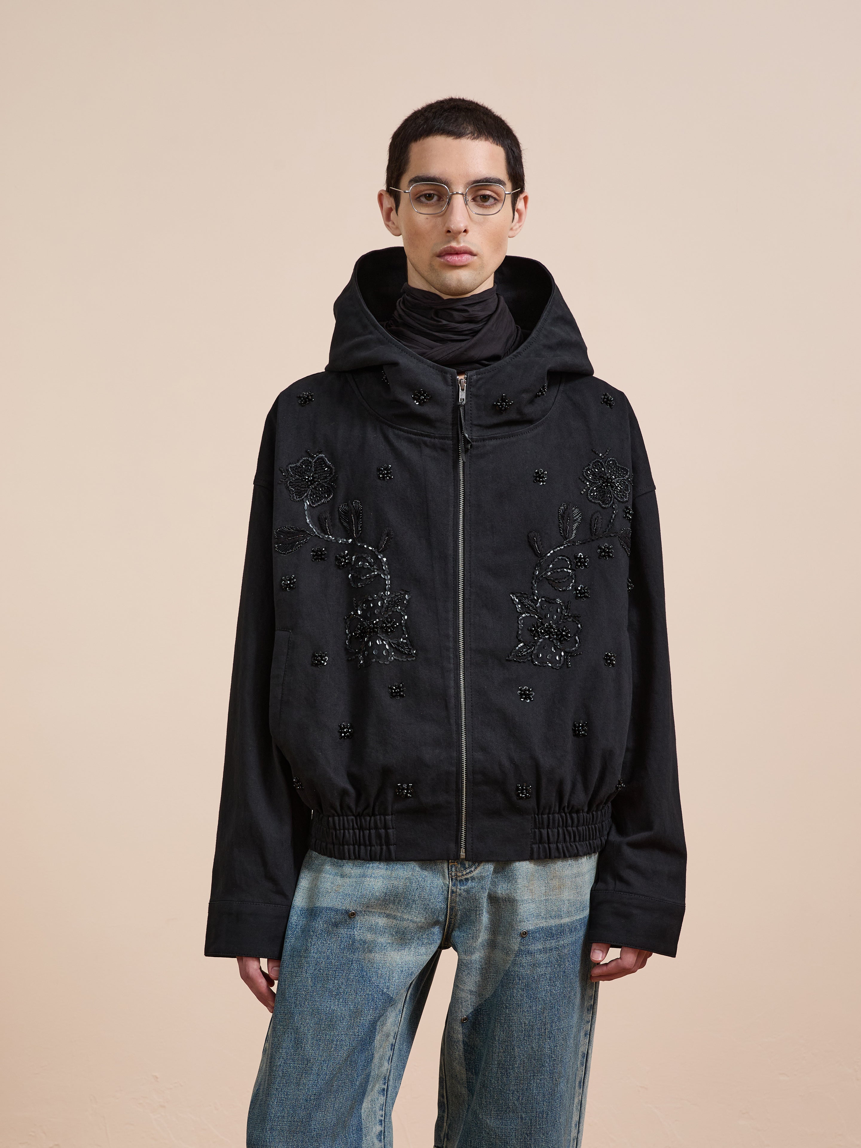 A person wearing glasses, a black FOUND Hand Embroidered Tonal Anorak Jacket with floral beadwork, and blue jeans stands against a beige background.