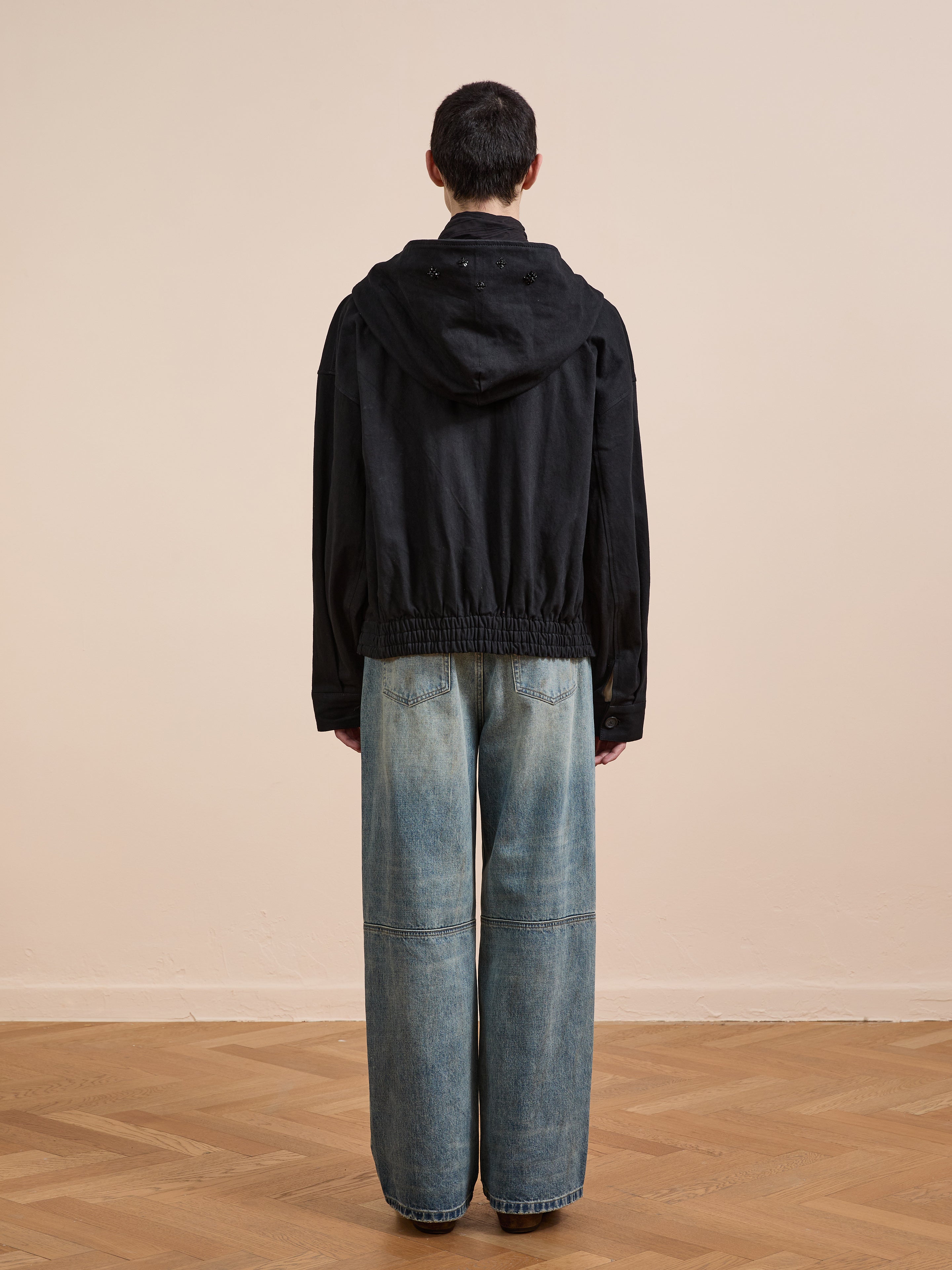 A person stands indoors on a wooden floor facing away, wearing FOUND's Hand Embroidered Tonal Anorak Jacket paired with wide-leg blue jeans.