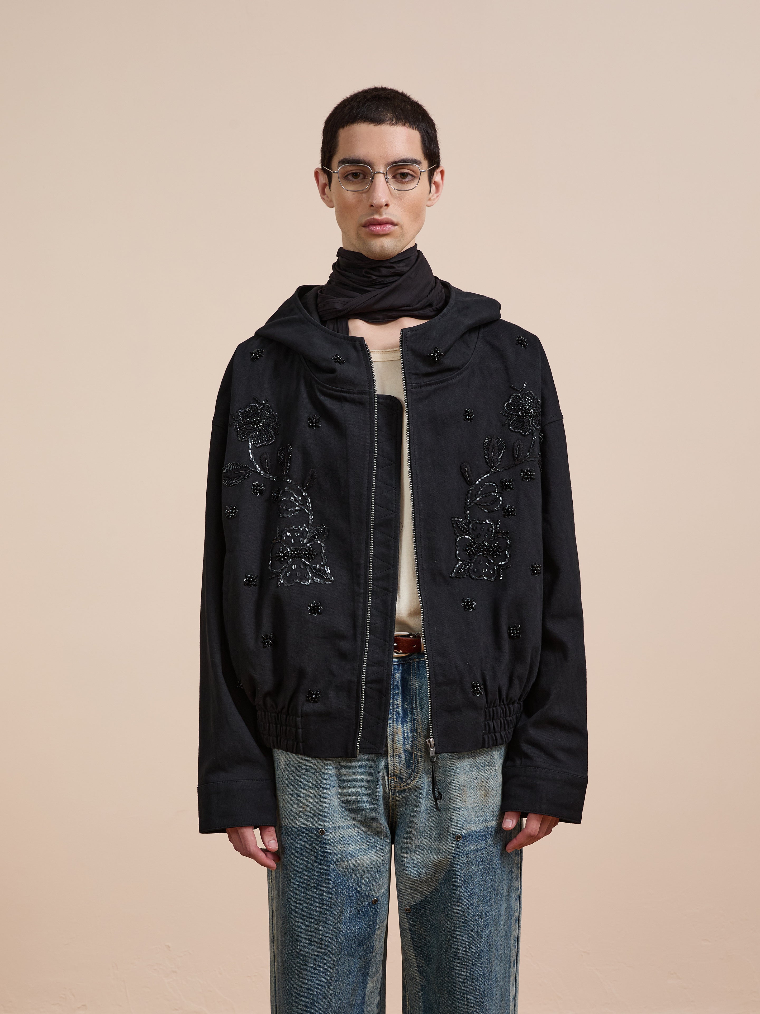 A person with short hair and glasses wears the FOUND Hand Embroidered Tonal Anorak Jacket over a light shirt and blue jeans, standing against a plain beige background.