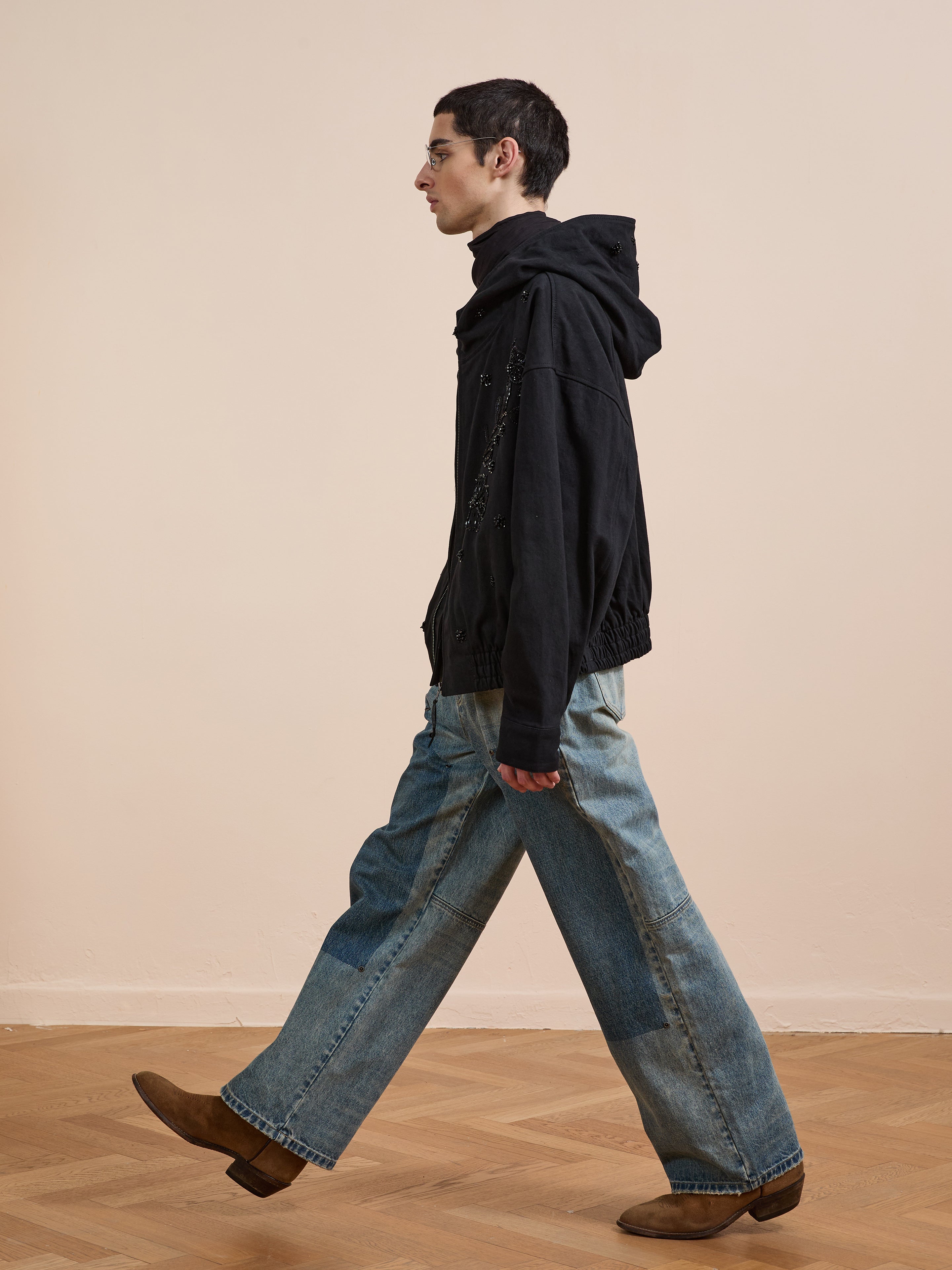 Wearing a black hoodie, wide-leg patchwork jeans, and brown boots, a person walks indoors on a wooden floor by a beige wall, layering with the FOUND Hand Embroidered Tonal Anorak Jacket for an artistic accent.