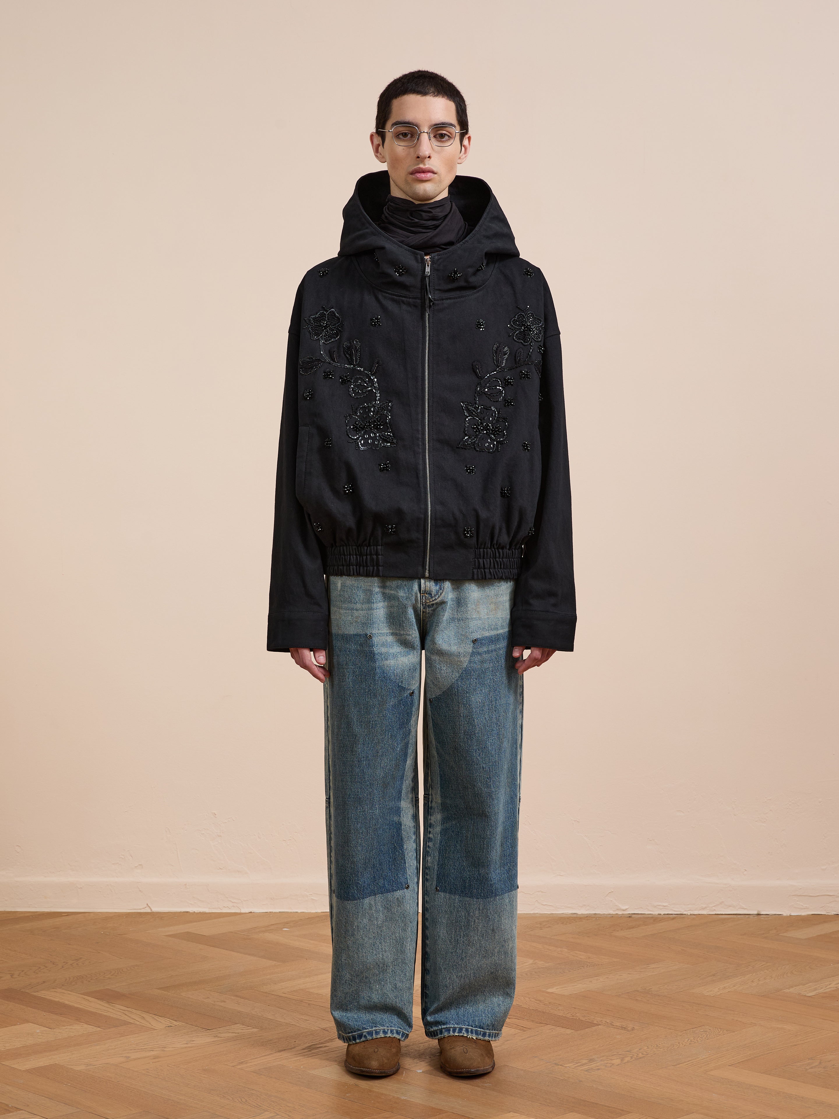 A person stands indoors wearing glasses, a FOUND Hand Embroidered Tonal Anorak Jacket, patchwork blue jeans, and brown shoes on a light wooden floor against a beige wall.