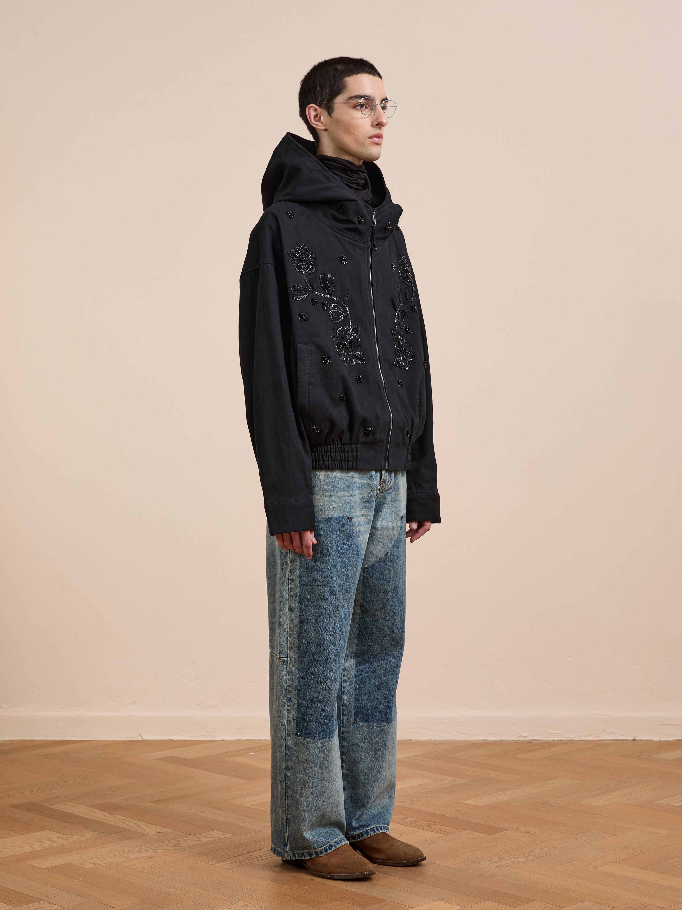 Indoors on a wooden floor, a person with glasses wears the FOUND Hand Embroidered Tonal Anorak Jacket, loose blue jeans with patches, and brown shoes.