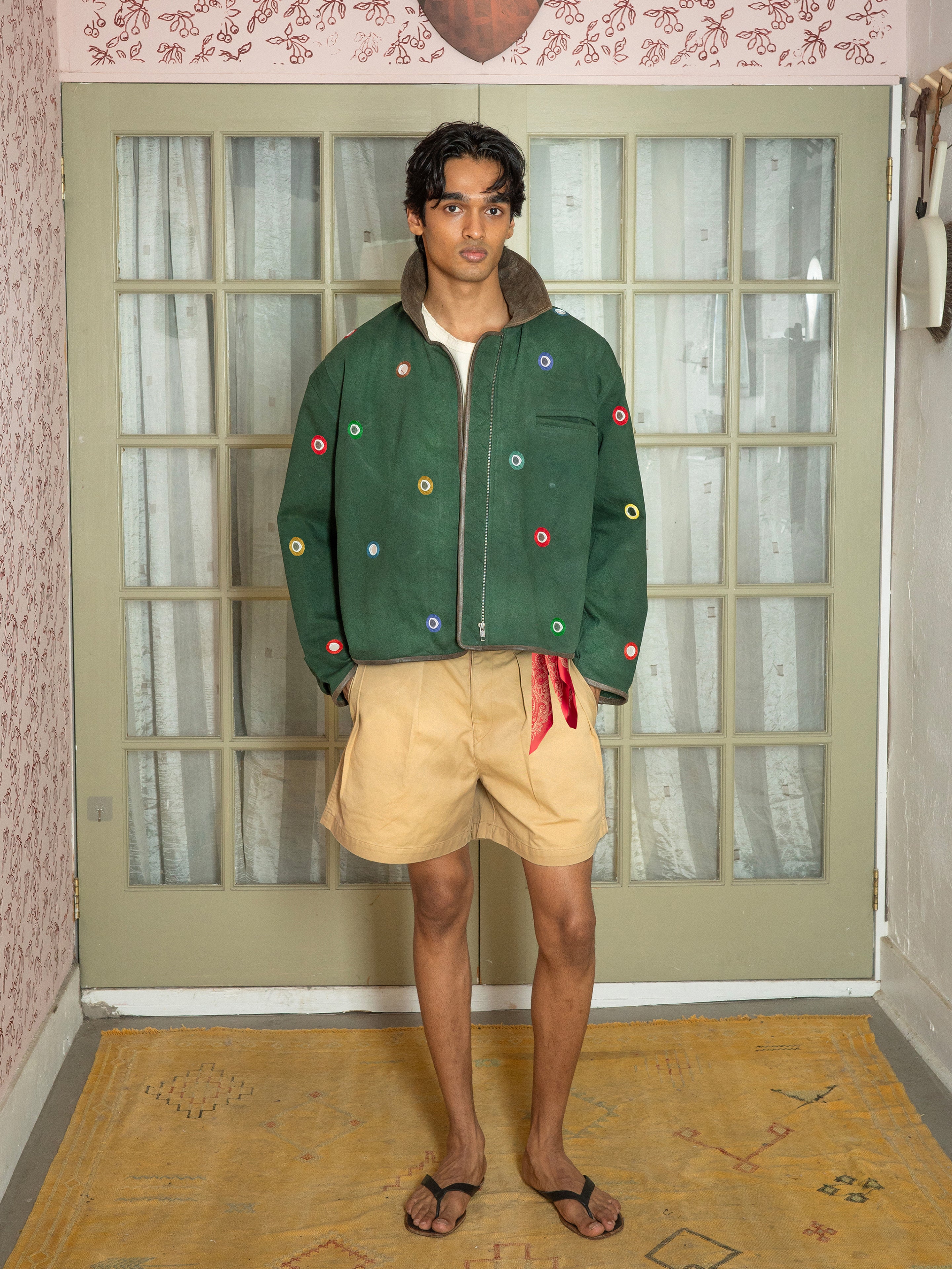 Indoors, a person wears the Found Hand Embroidered Mirror Abhala Jacket with colorful circular accents, tan shorts, a red bandana tucked in the waistband, and black sandals.