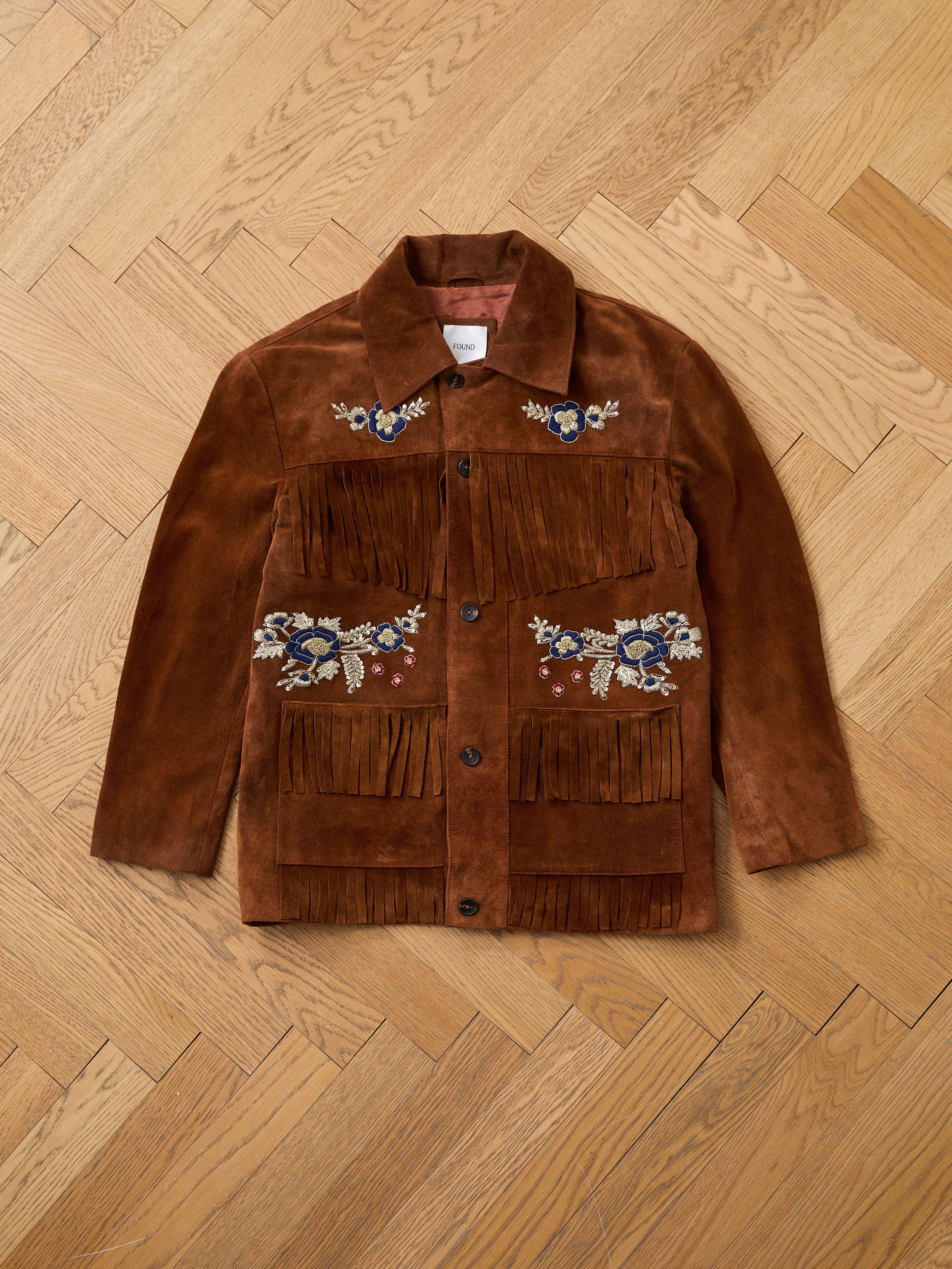 The FOUND Hand Embroidered Suede Fringe Jacket in brown, featuring colorful floral designs, is laid flat on a light wood herringbone floor.