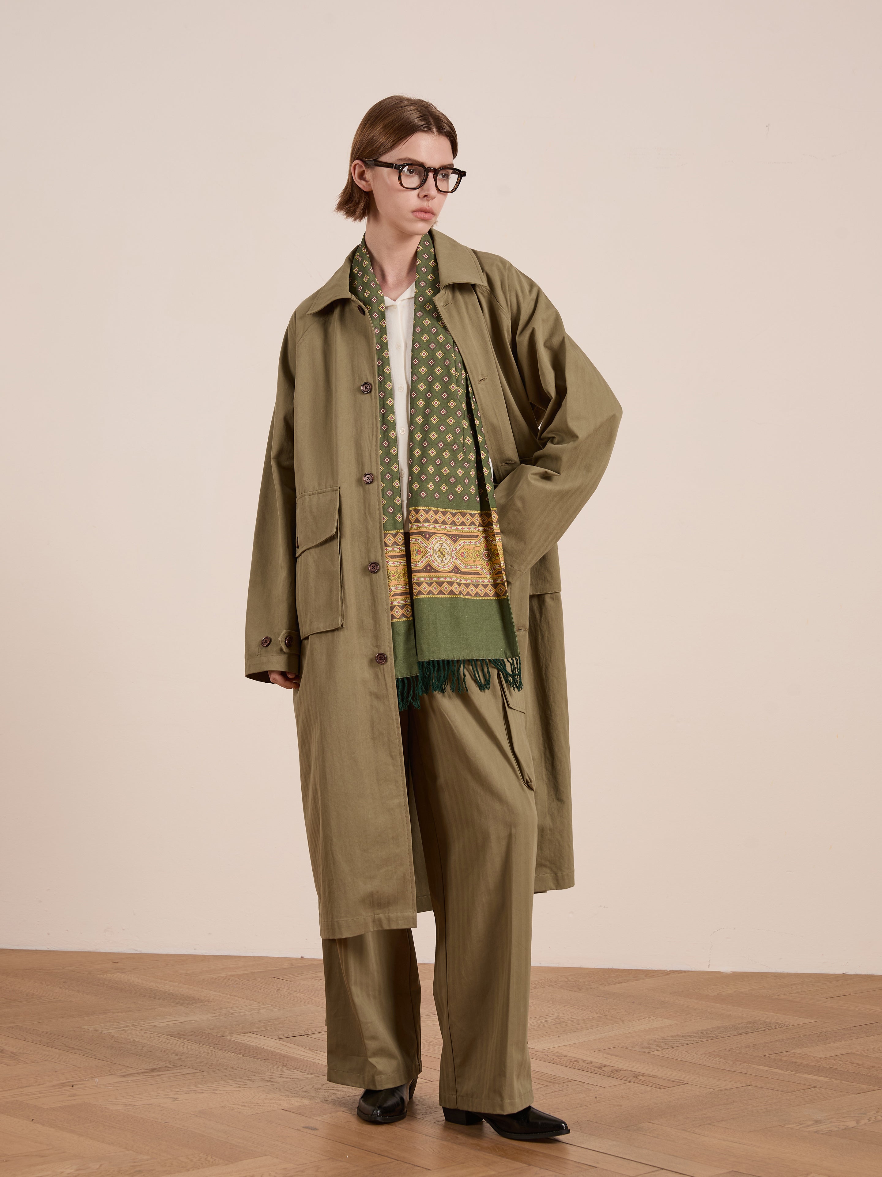 A person stylishly dons a FOUND Herringbone Twill Trench Coat, a patterned scarf, and glasses in a room with wooden flooring. Brown shoes complete the chic unisex look, elevating the casual yet sophisticated vibe.