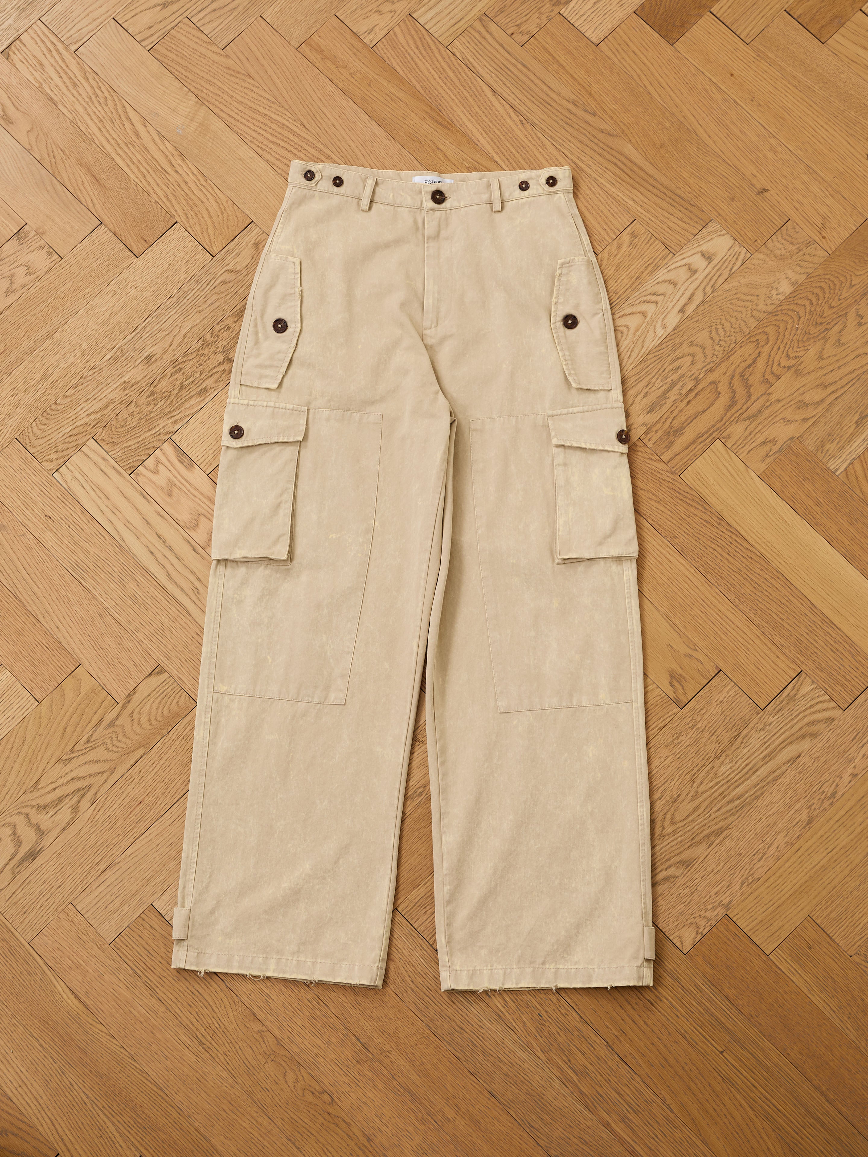 FOUND Utility Herringbone Paneled Pants in beige, featuring a unisex relaxed fit, side pockets, and button closures, shown laid flat on a wooden parquet floor.