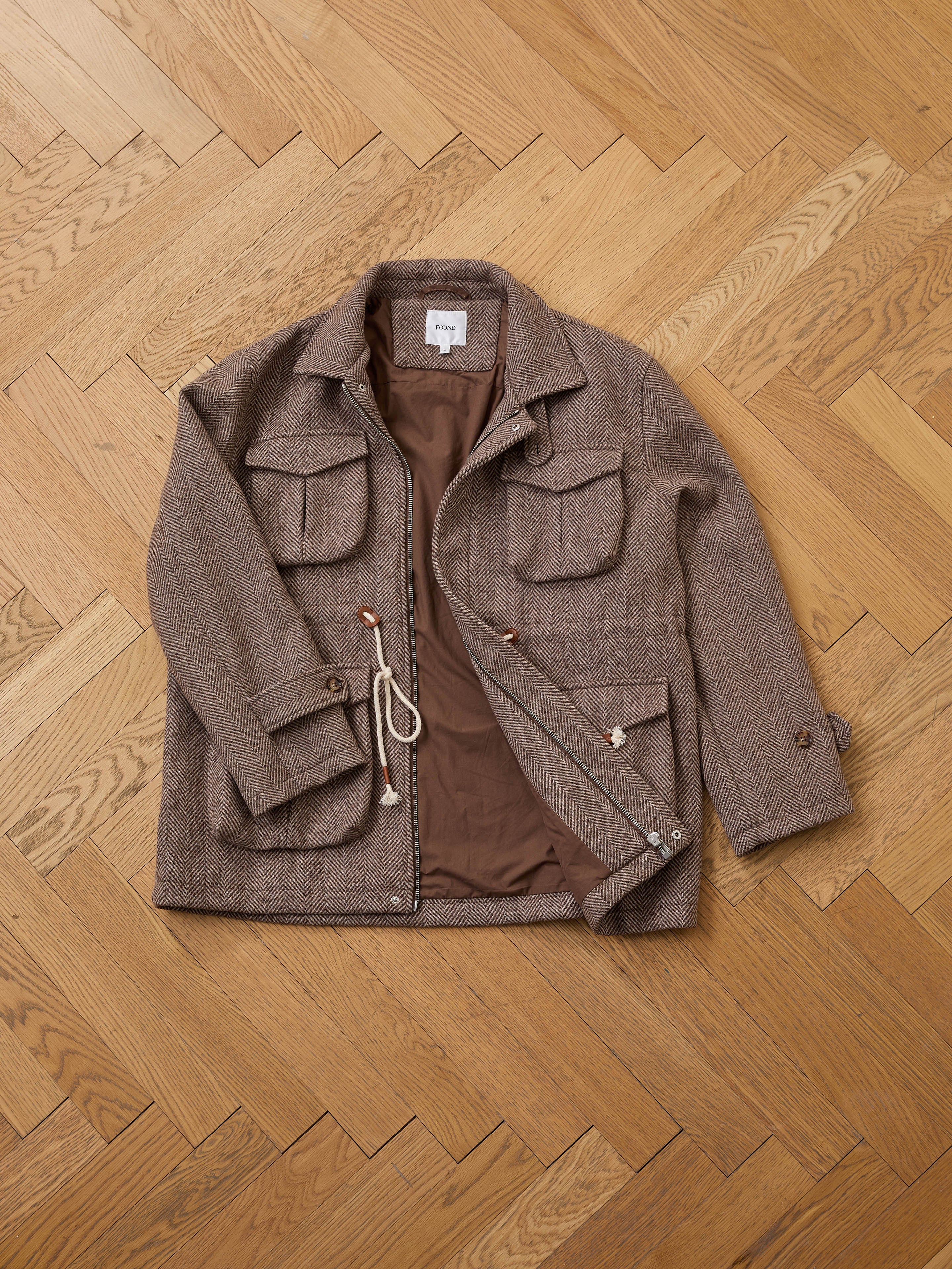 The FOUND Herringbone Wool Barn Jacket, featuring a brown wool blend, four front pockets, button and zip closures, and a rope drawstring, is laid flat on a light wood herringbone floor.
