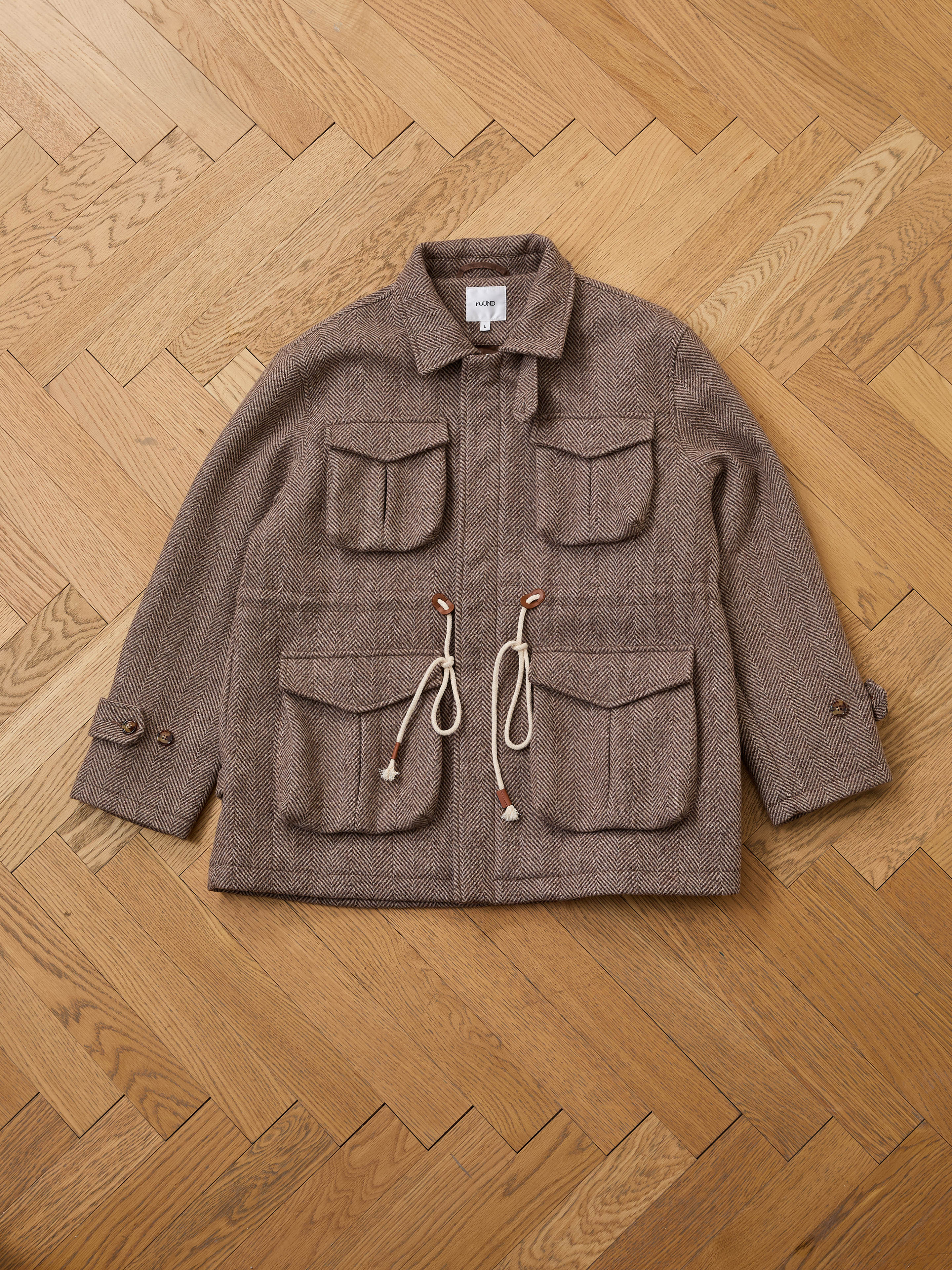 The FOUND Herringbone Wool Barn Jacket, featuring a brown wool blend, four front pockets, buttoned cuffs, collar, and drawstring waist, is laid flat on a herringbone wooden floor.