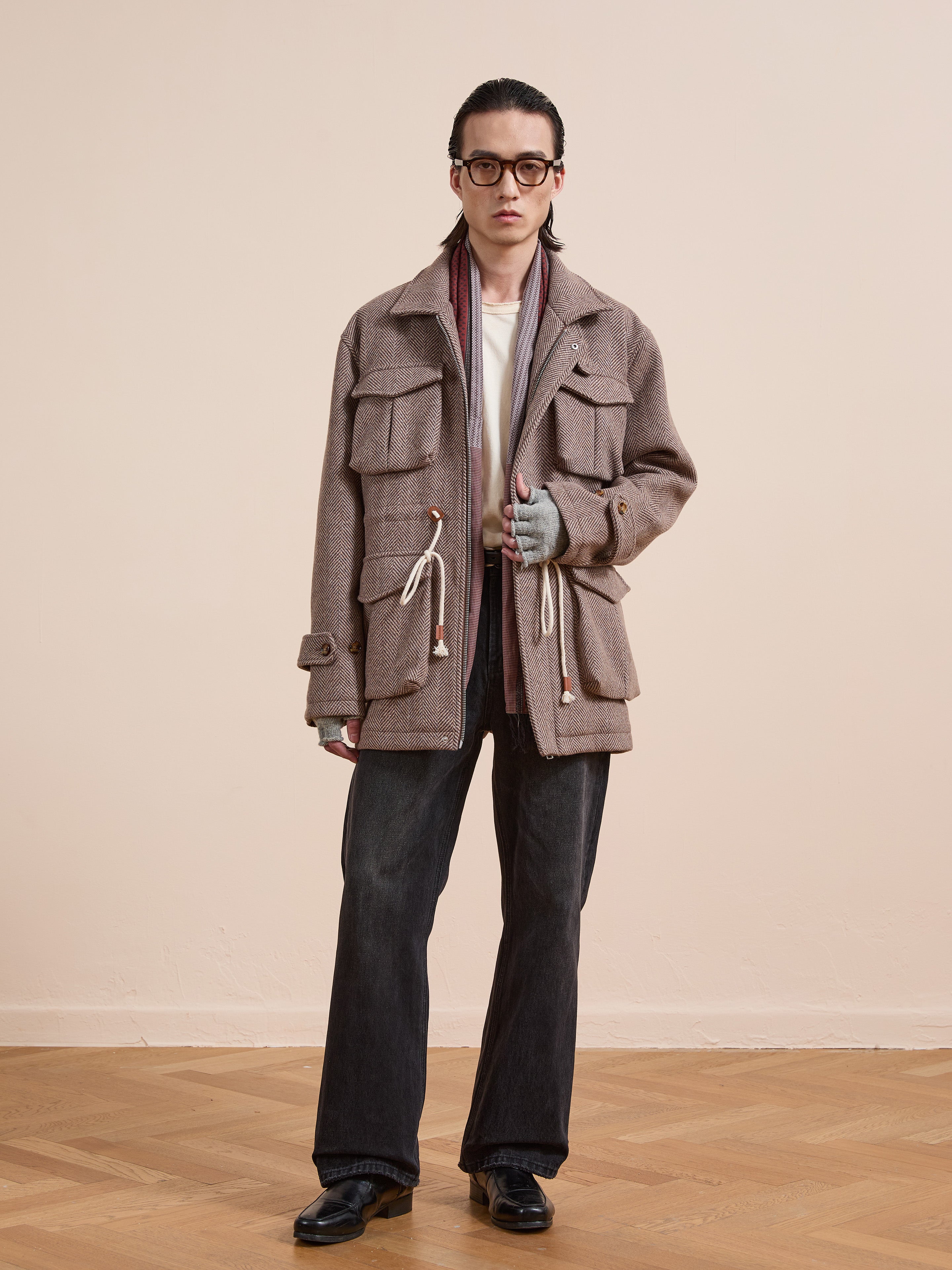 A person wearing glasses, a FOUND Herringbone Wool Barn Jacket with large pockets, a scarf, fingerless gloves, black jeans, and black shoes stands indoors against a plain background.