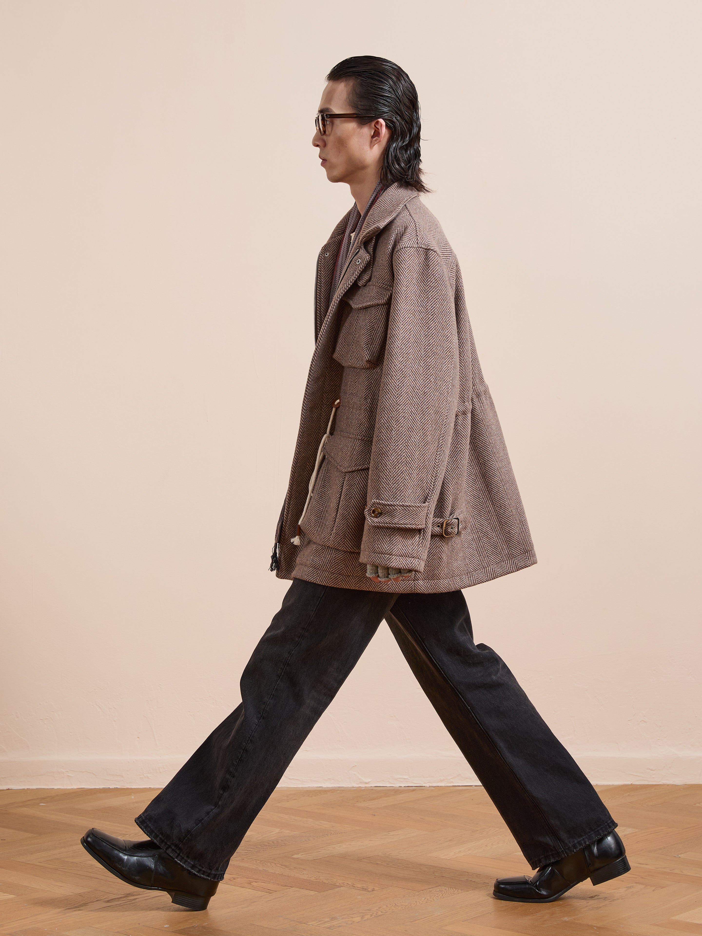A person with slicked-back hair walks in profile wearing the FOUND Herringbone Wool Barn Jacket, black bootcut pants, black shoes, and sunglasses on a wooden floor against a plain background.