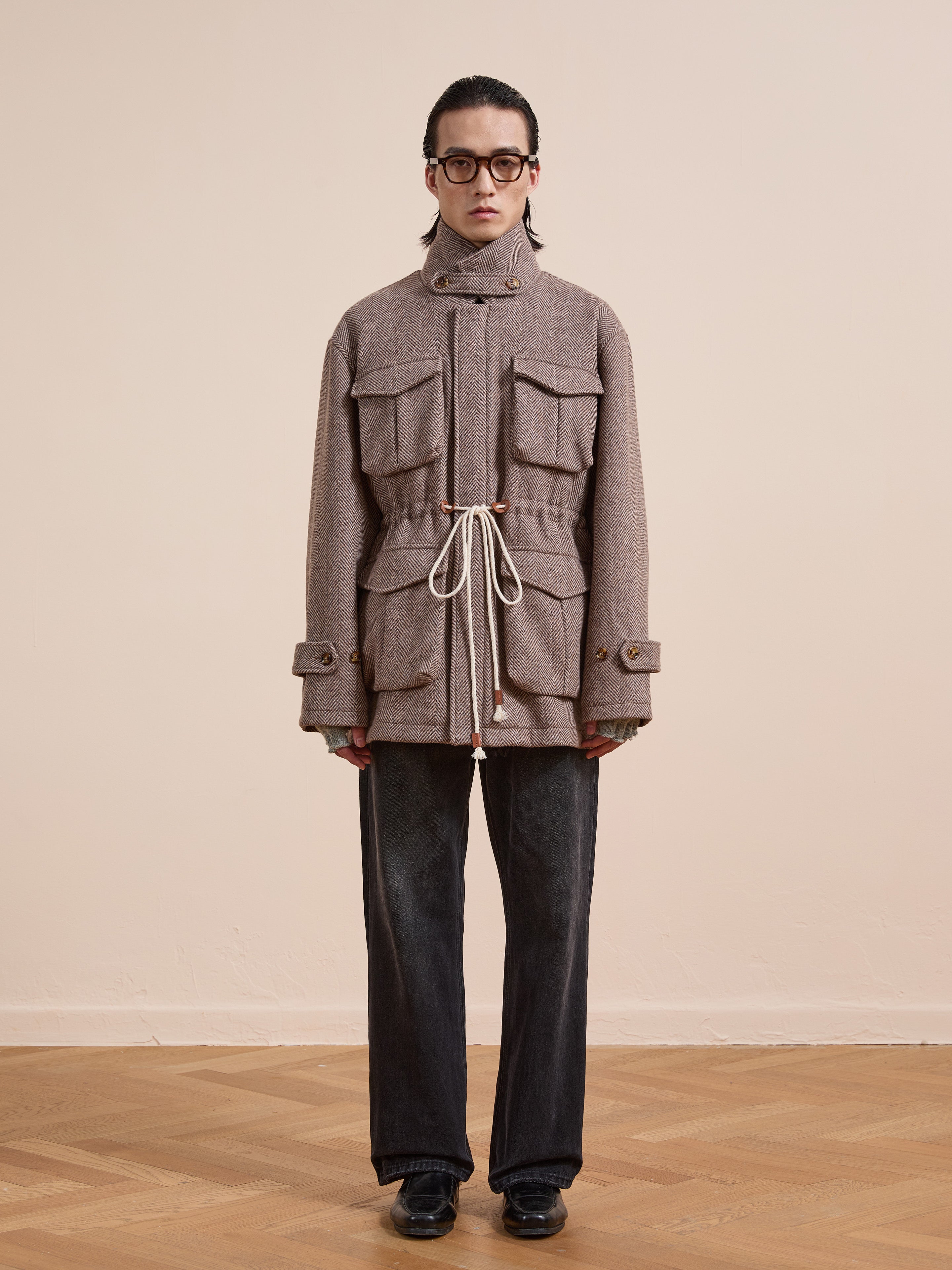 A person stands indoors against a plain wall, wearing glasses and the FOUND Herringbone Wool Barn Jacket with large pockets and a drawstring waist, paired with dark pants and black shoes.