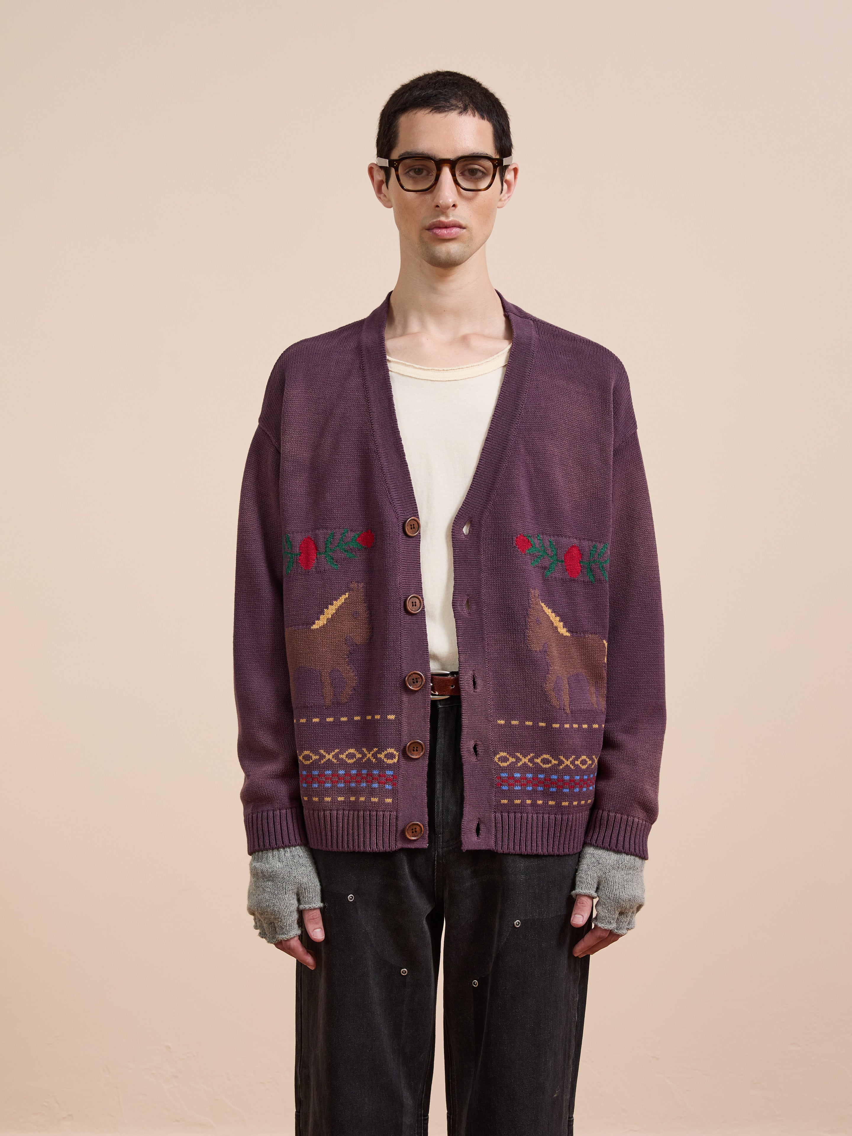 A person in glasses, a cream shirt, black pants, fingerless gloves, and the Found Horse Jacquard Cardigan featuring animal and plant patterns stands against a plain beige background.
