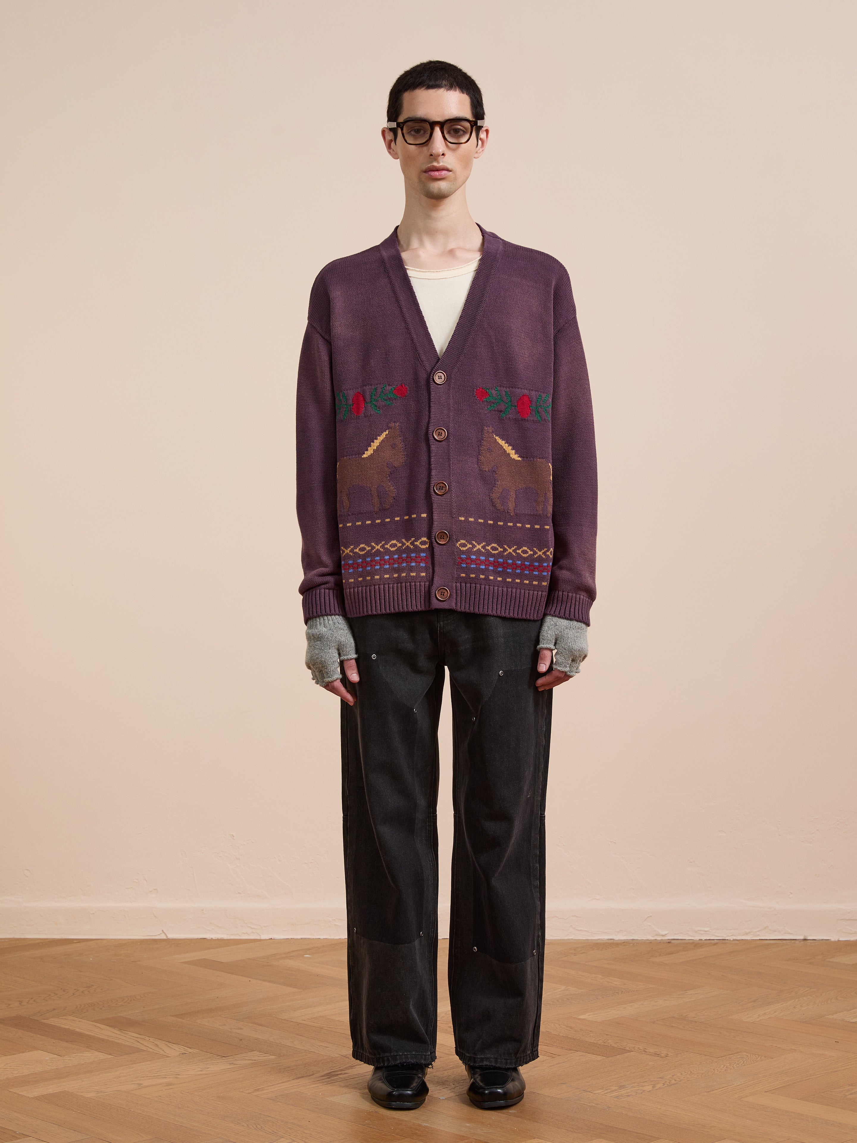 Indoors, a person wears Found’s Horse Jacquard Cardigan, featuring unisex equestrian-inspired knit with deer and geometric patterns, paired with glasses, a white shirt, gray fingerless gloves, black pants and black shoes.