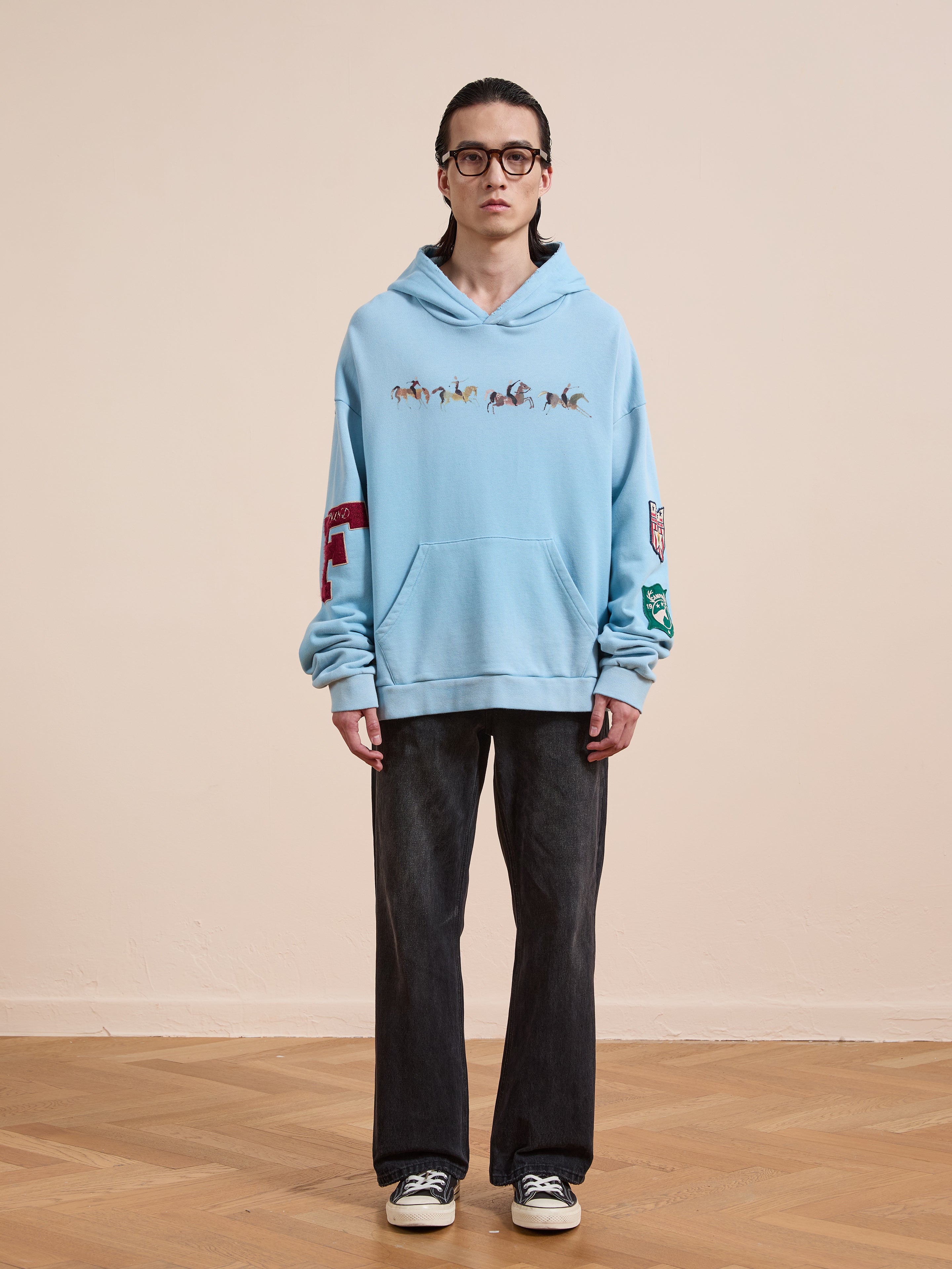 A person wearing glasses, a light blue FOUND Horse Line Hoodie in soft French Terry cotton with small horse graphics, dark jeans, and black sneakers stands on a wooden floor against a plain beige wall.