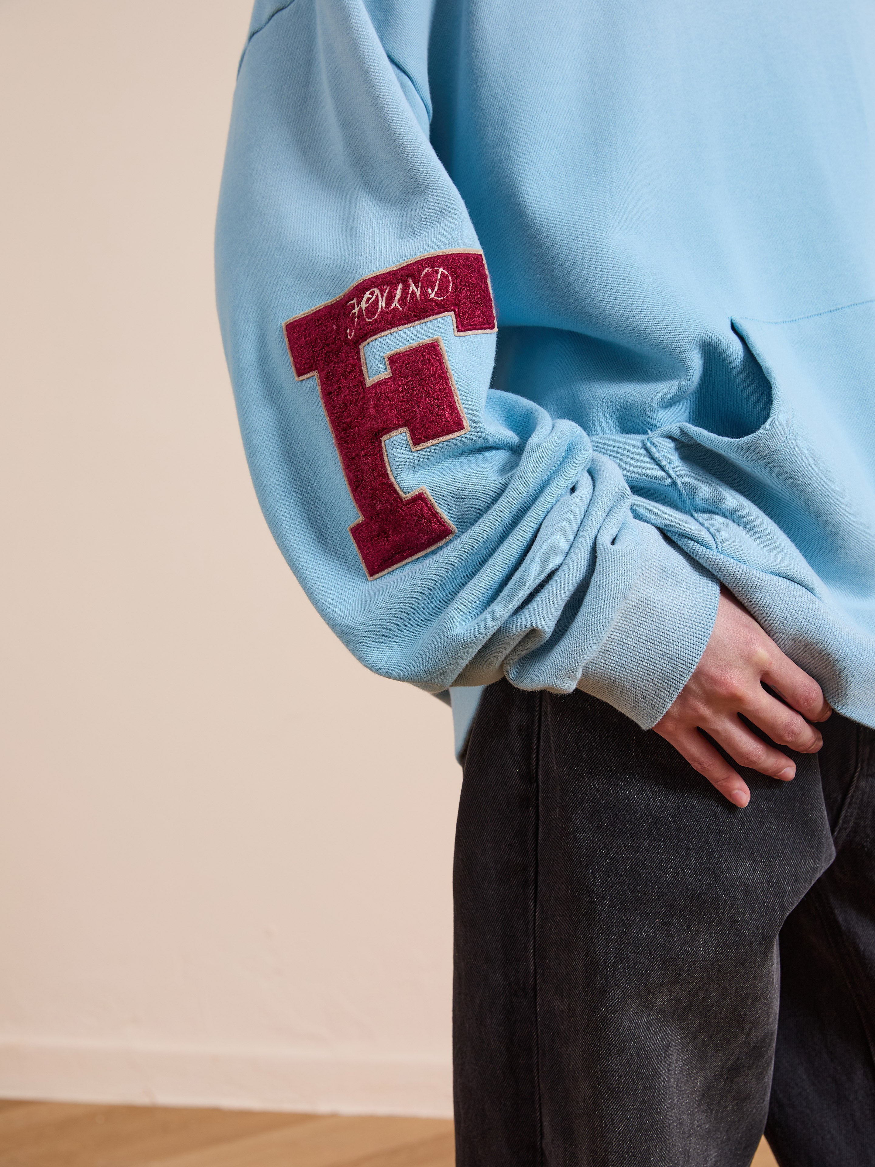 A person wearing the FOUND Horse Line Hoodie—light blue, vintage style, soft French Terry cotton with a red "F" patch on the sleeve—and dark jeans stands against a plain background.