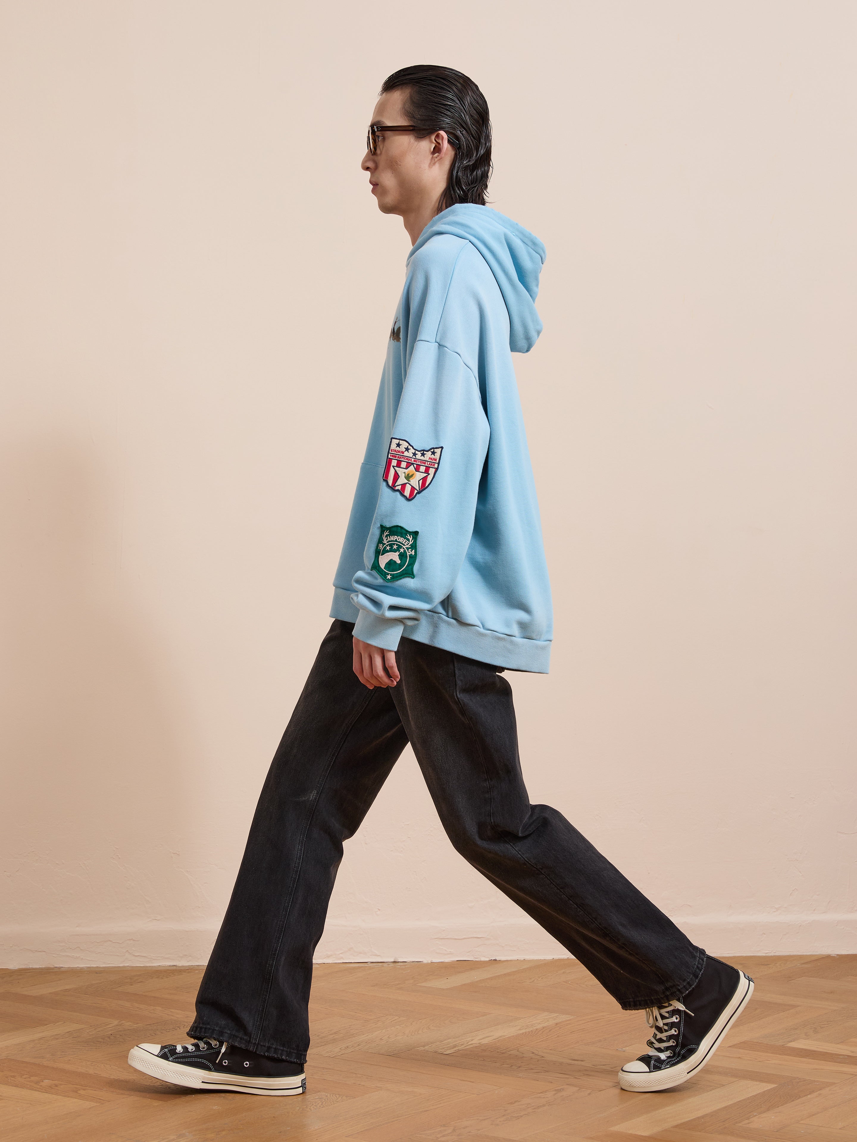 A person in sunglasses, black wide-leg jeans, and black sneakers walks indoors on a wooden floor wearing the FOUND Horse Line Hoodie in light blue French Terry cotton with patches.