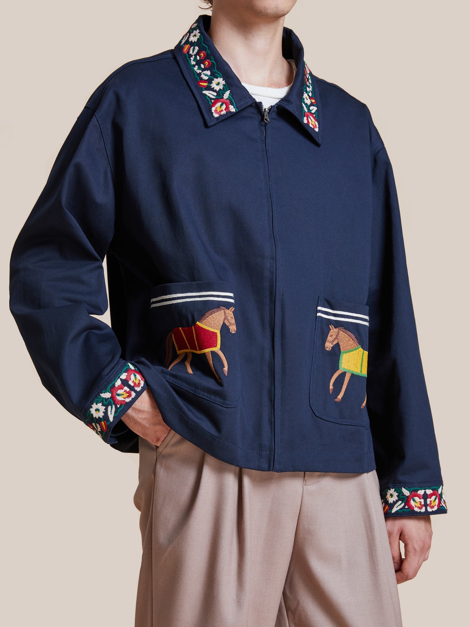 Man wearing a dark blue Found Horse Equine Work Jacket with detailed embroidery on the sleeves and floral patterns on the collar and cuffs, paired with light brown trousers.