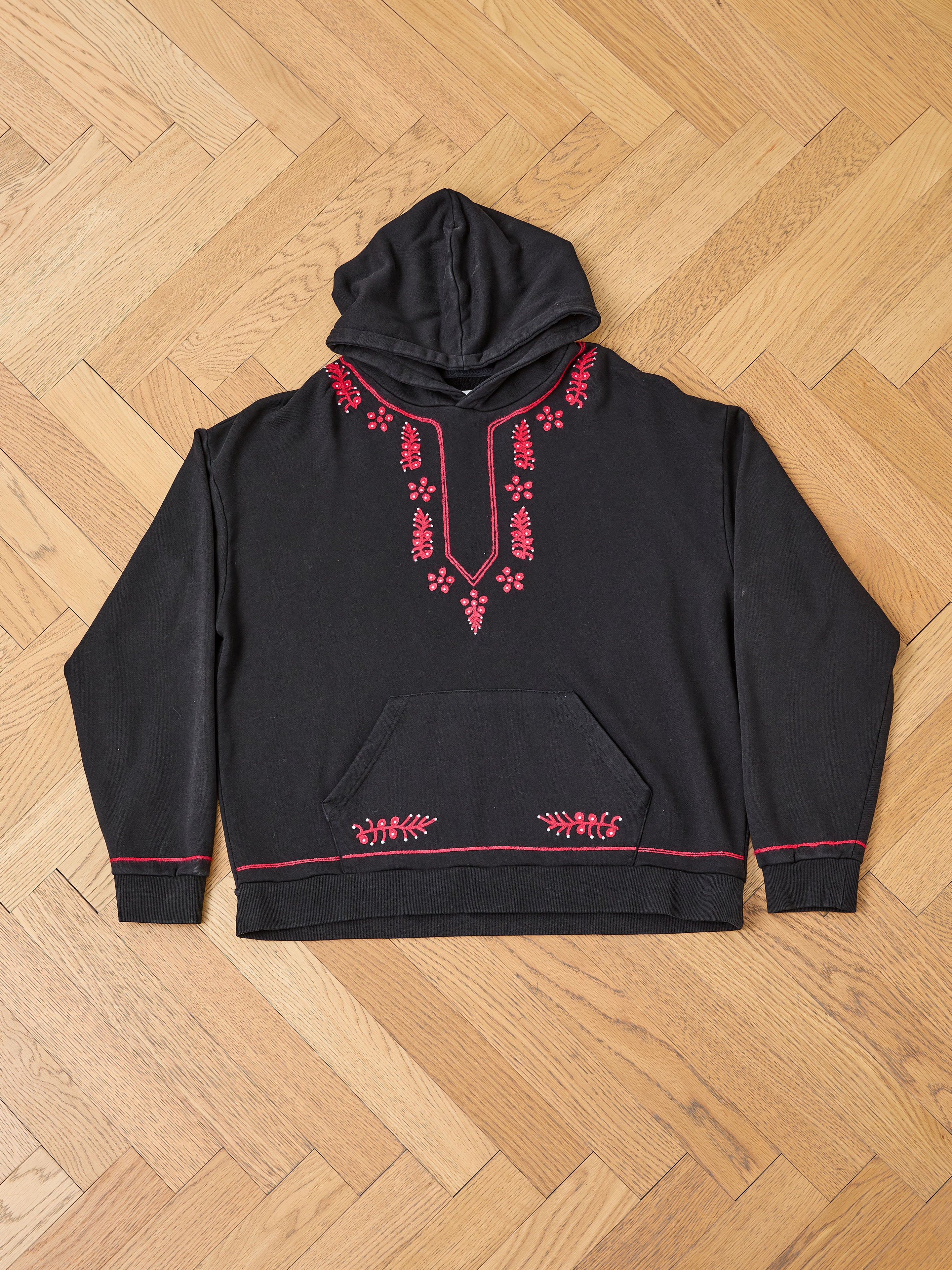The FOUND Indus Embroidered Hoodie, black with red floral embroidery on the chest, neckline, and pocket, is shown laid flat on a wooden parquet floor.