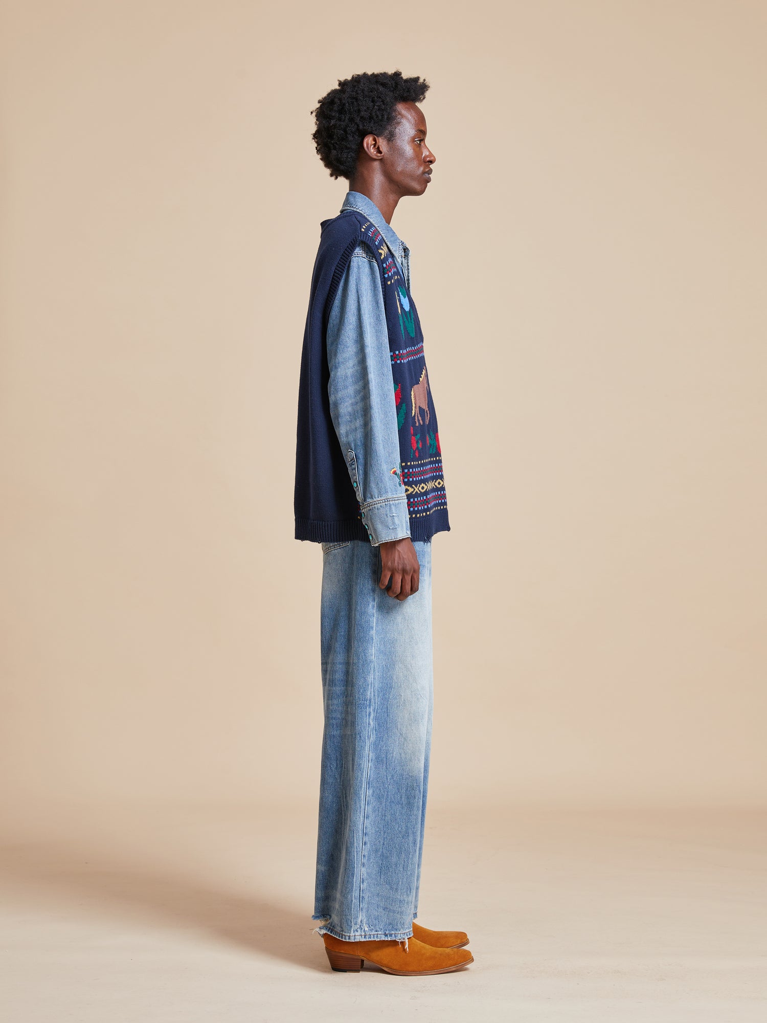 A man wearing the Found Isle Horse Reverie Sweater Knit Vest and jeans standing in front of a beige background.