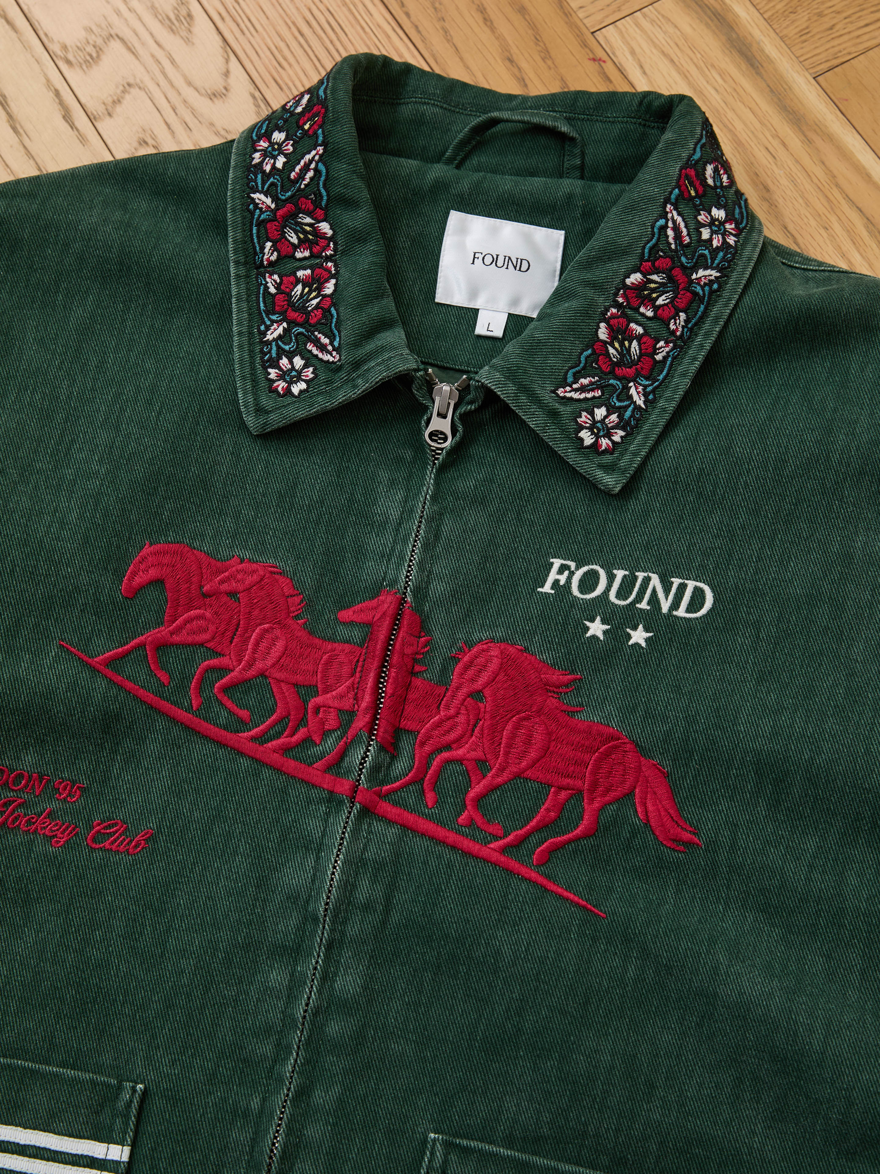 The Found Jockey Club Embroidered Jacket features red racing horses, floral collar details, and "FOUND" with two stars on the chest. This unisex relaxed fit jacket is shown laid flat on a wooden floor.