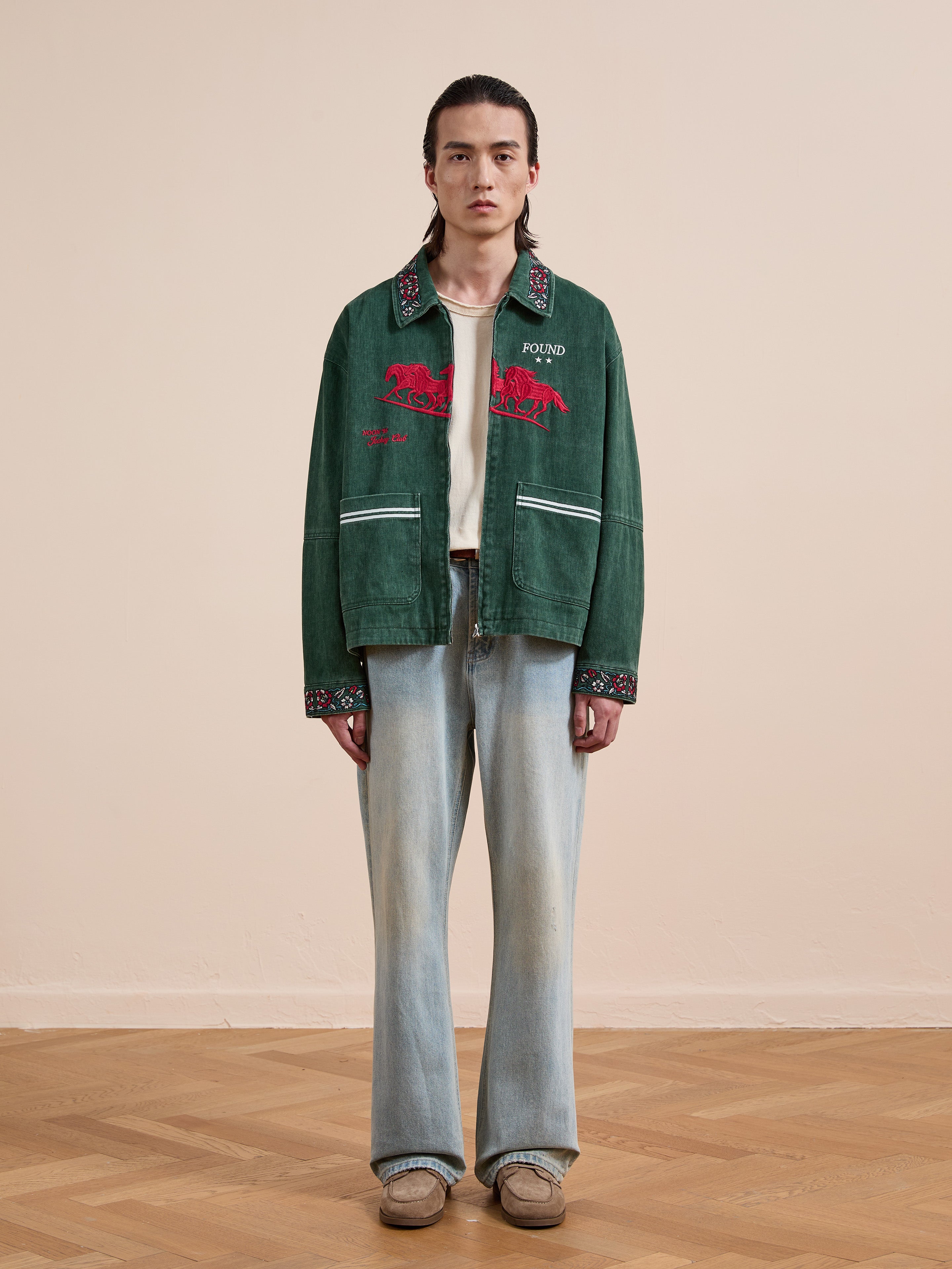 A person stands indoors on a wooden floor against a beige wall, wearing the Found Jockey Club Embroidered Jacket in green over a white shirt, faded blue jeans, and brown shoes.