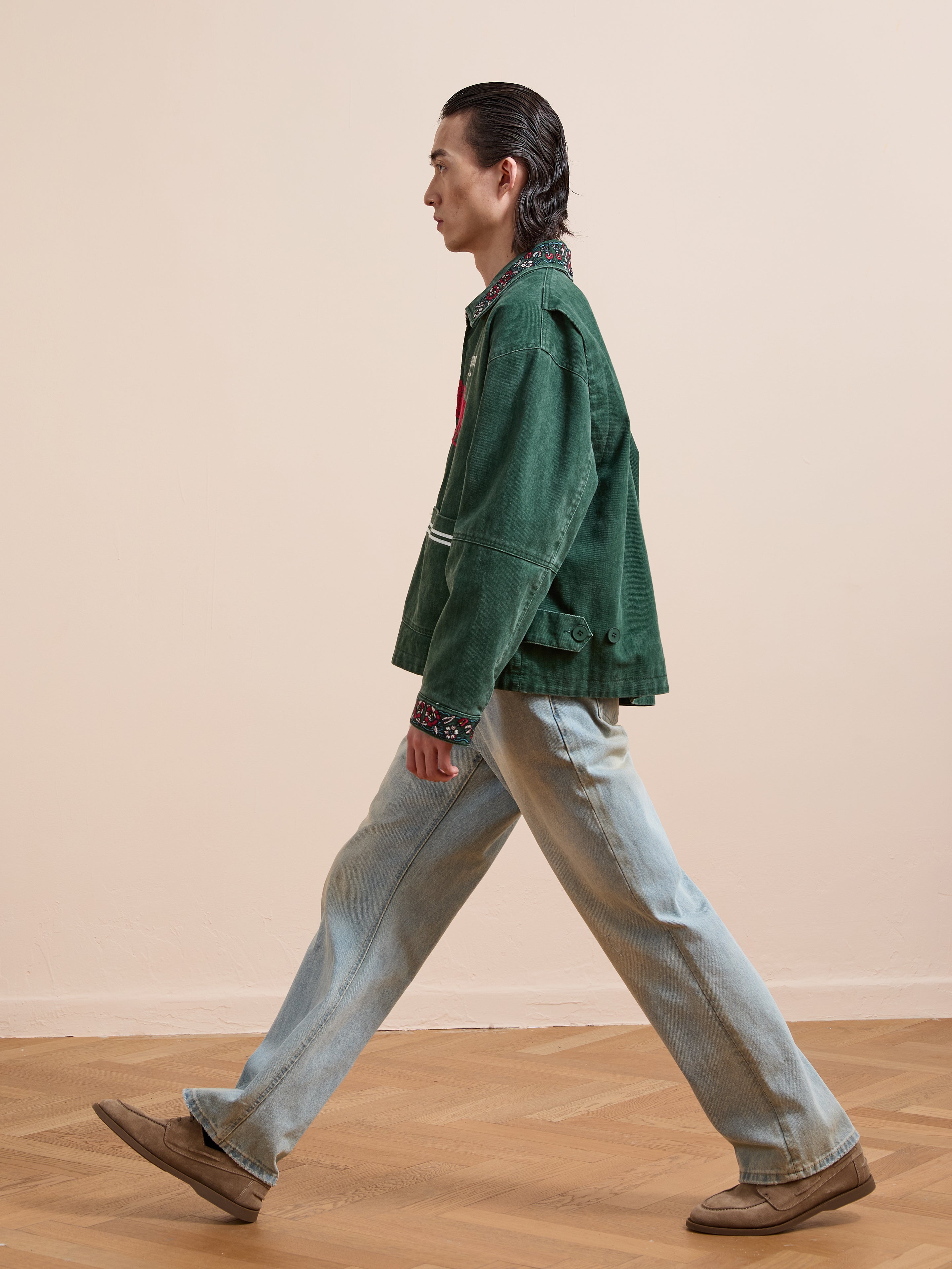 A person with slicked-back hair walks indoors, wearing a green Found Jockey Club Embroidered Jacket with racing horses, light blue jeans, a patterned shirt, and brown shoes.