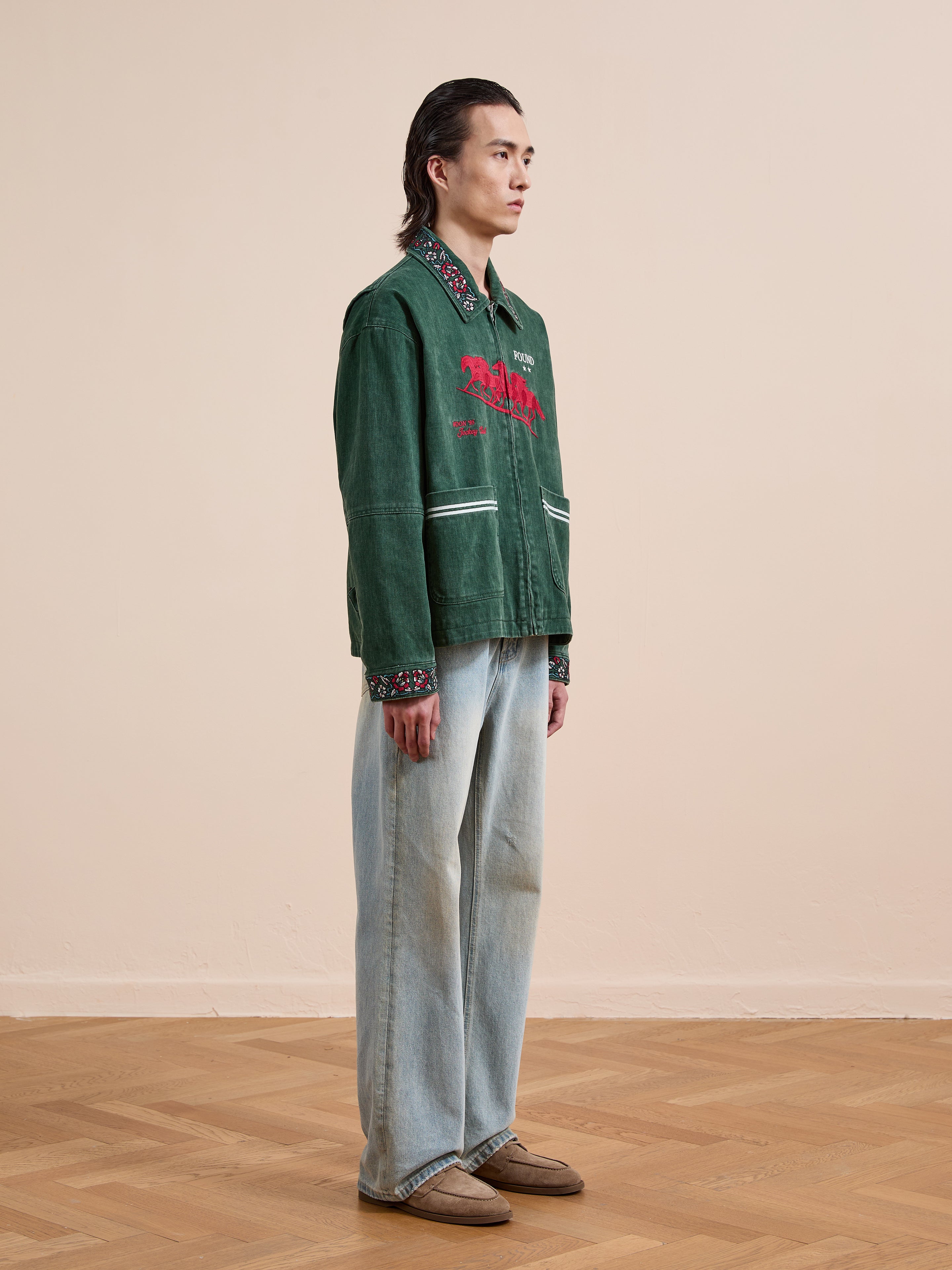 A person stands indoors on a wooden floor, wearing the Found Jockey Club Embroidered Jacket, light blue wide-leg jeans, and brown shoes, facing sideways to the camera.