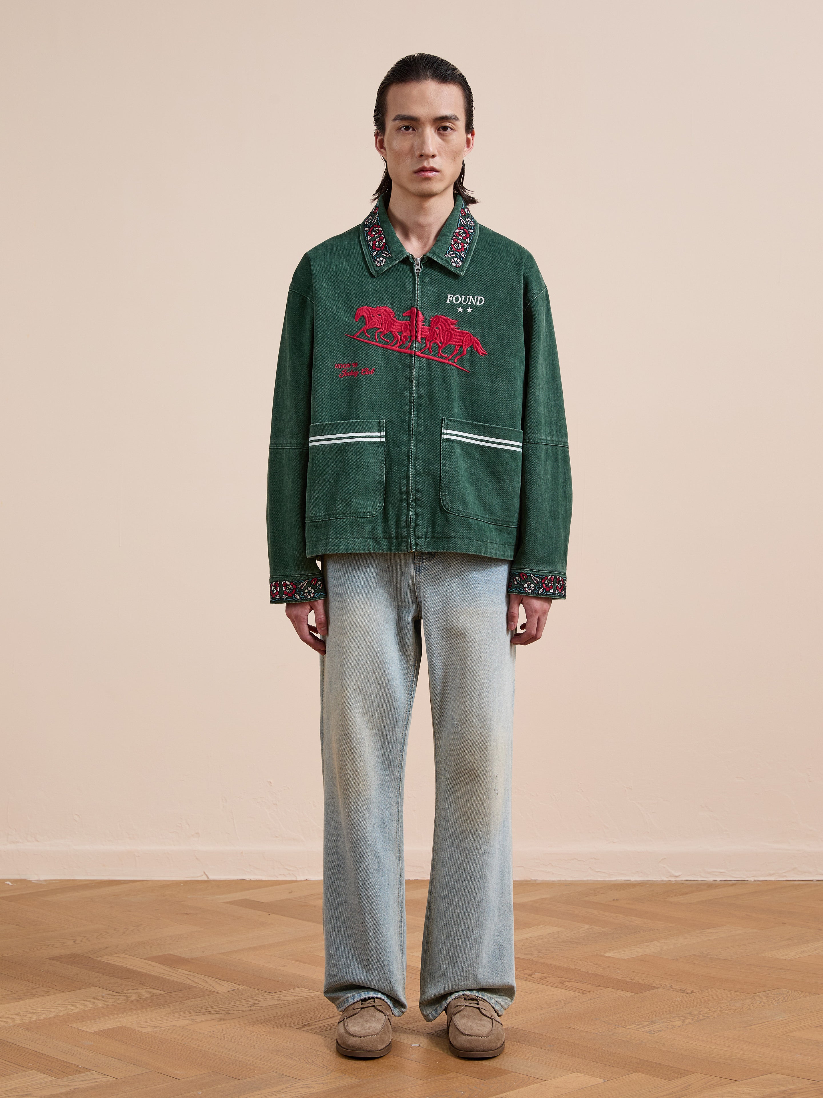 A person stands indoors against a light wall and wooden floor, wearing Found’s Jockey Club Embroidered Jacket in green with racing horses, styled with light blue jeans and beige shoes. The jacket offers a unisex relaxed fit.