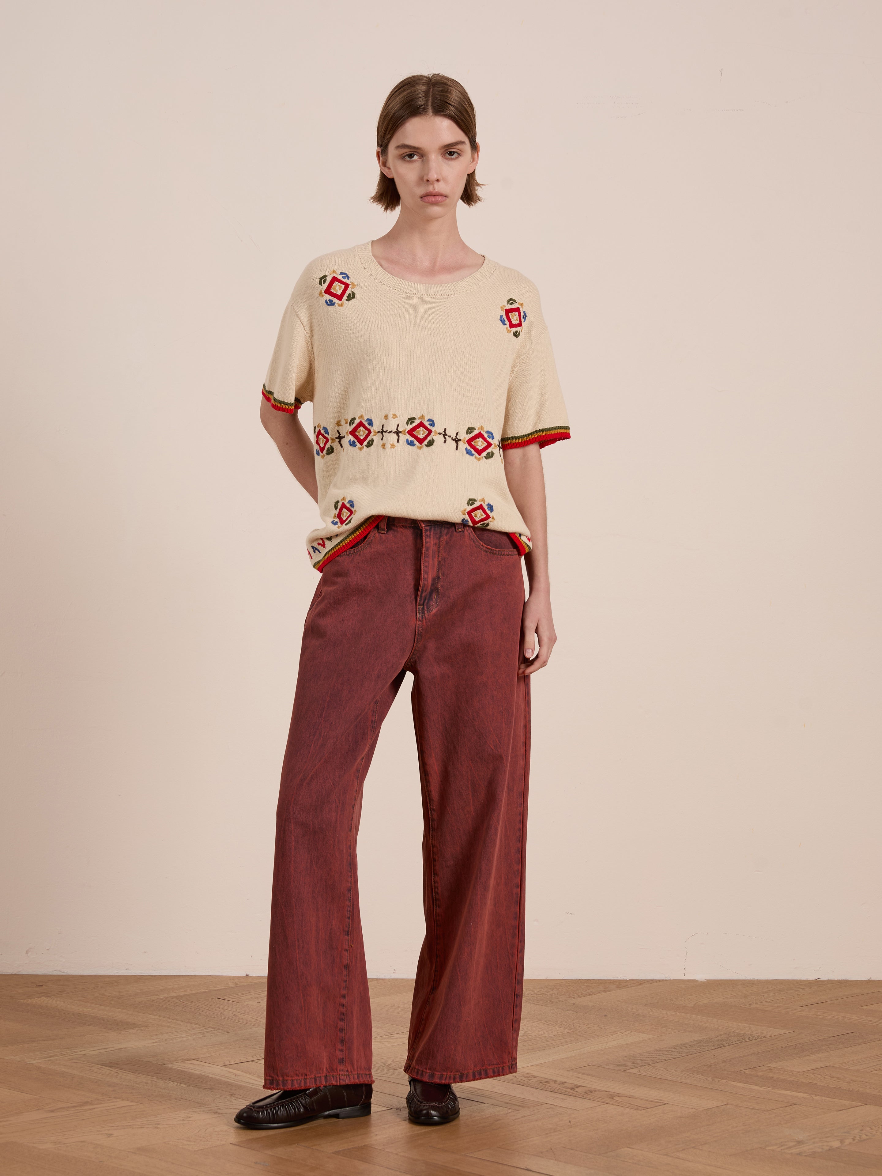 A person stands indoors against a plain wall on wooden flooring, wearing a vintage-inspired FOUND Knit Flower Petal Tee with colorful patterns and wide-legged maroon pants.