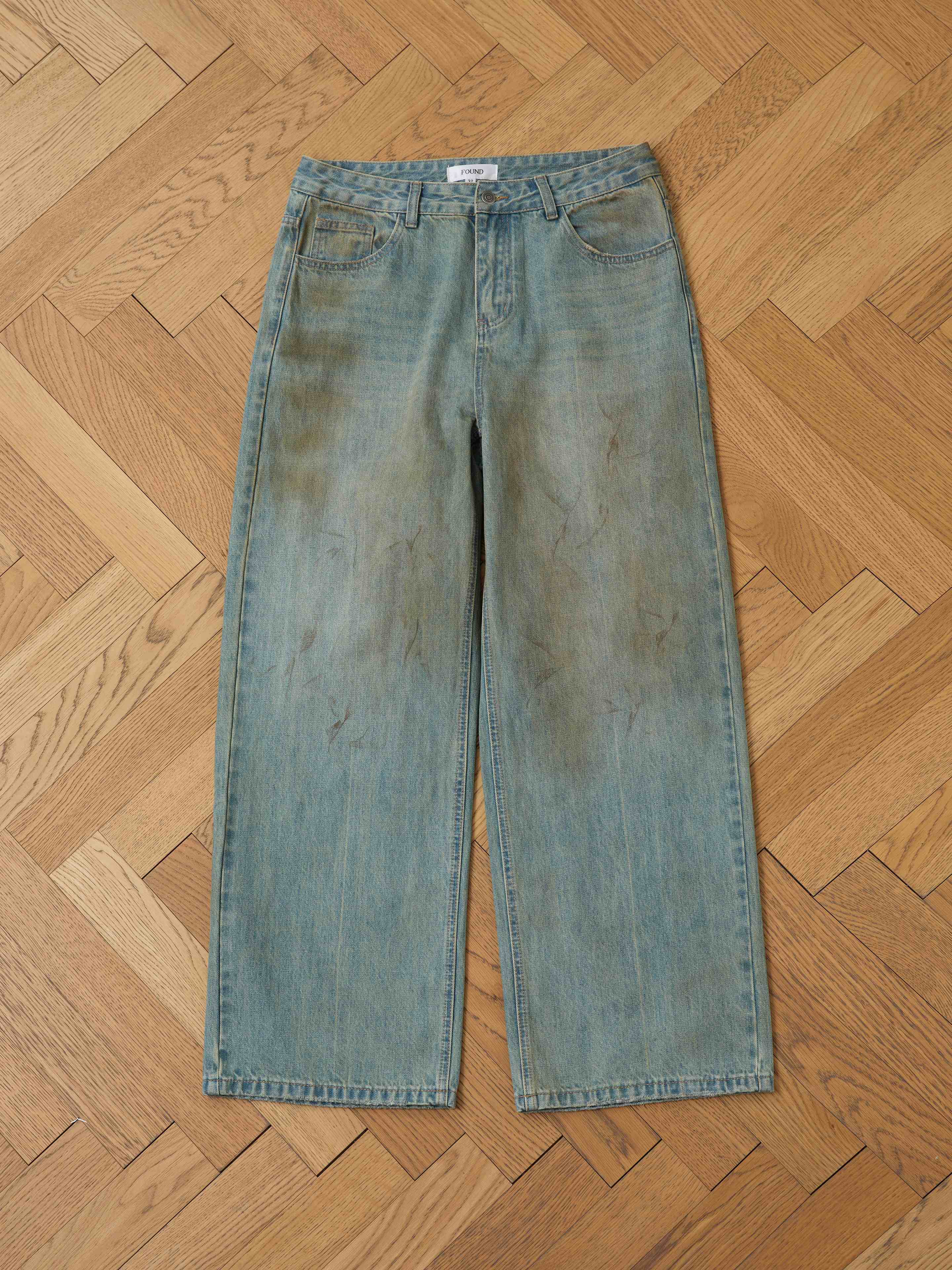 The FOUND Lacy Baggy Jeans, a unisex wide-leg style in dirty blue with visible stains and marks, are displayed flat on a wooden herringbone-patterned floor.