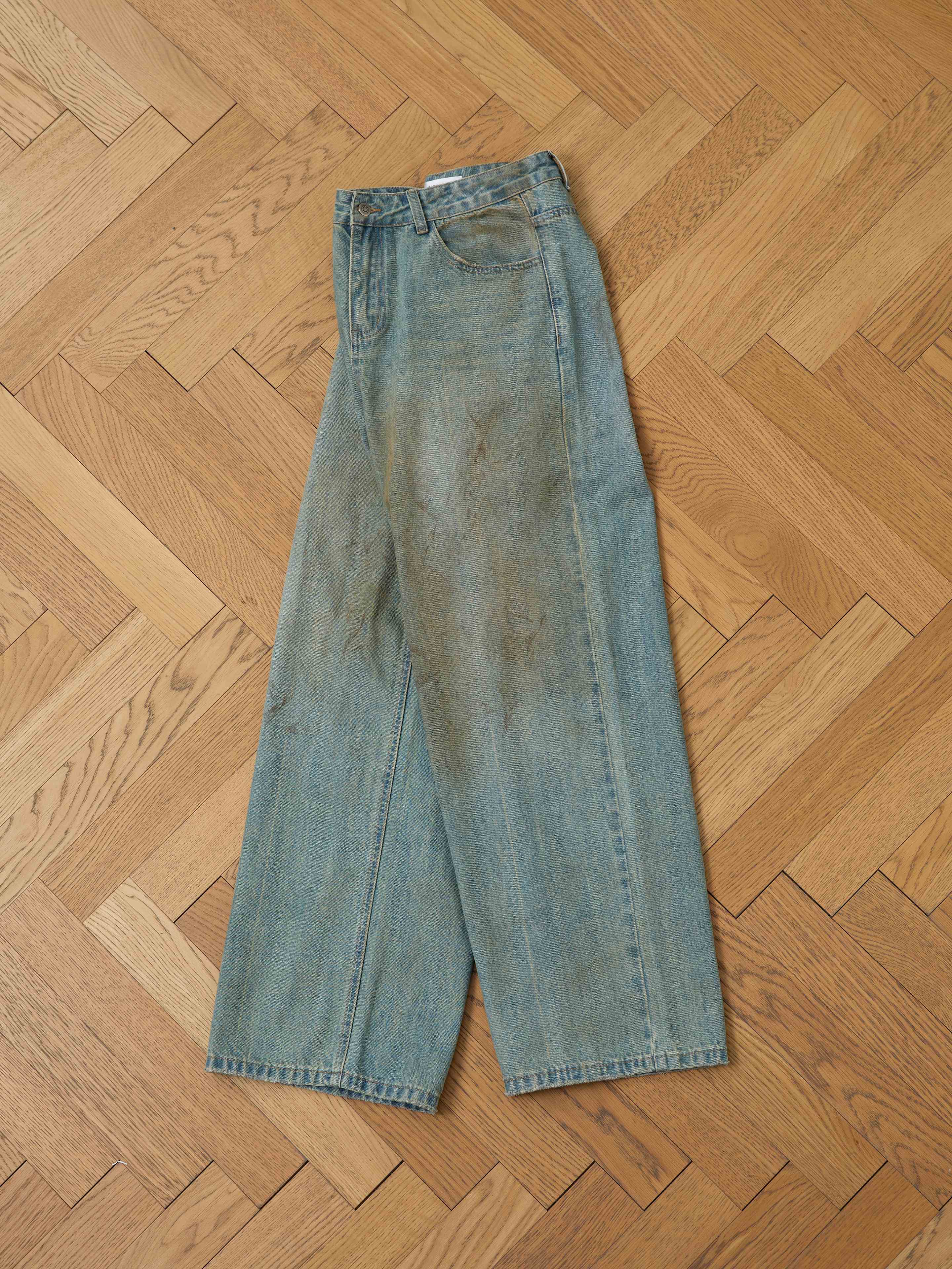 A pair of FOUND Lacy Baggy Jeans with a relaxed fit and visible stains is laid flat on a wooden parquet floor.