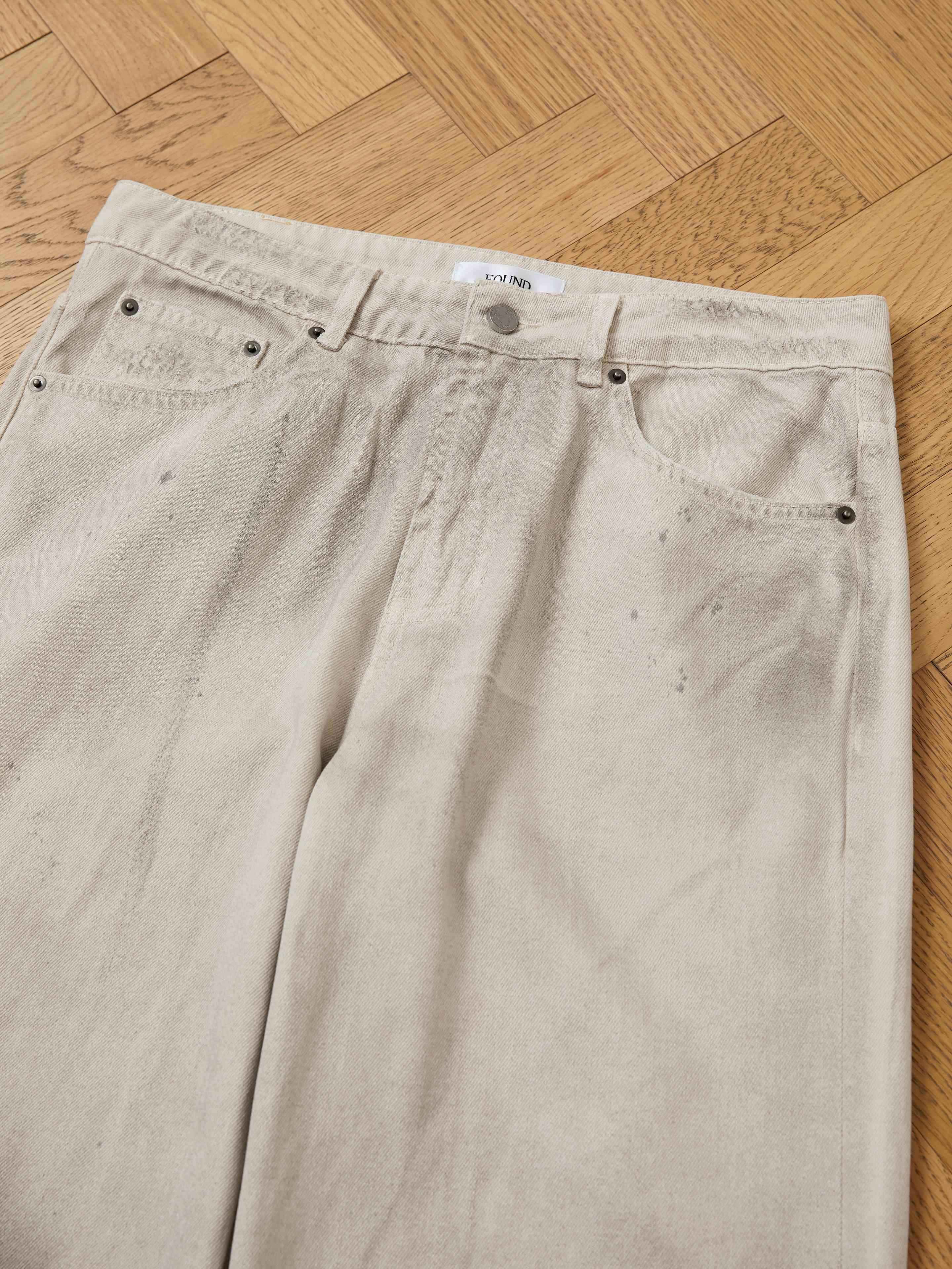 FOUND’s Lacy Baggy Jeans in a light wash feature a relaxed fit and subtle distressed detailing with faint stains, shown laid flat on a wooden parquet floor.