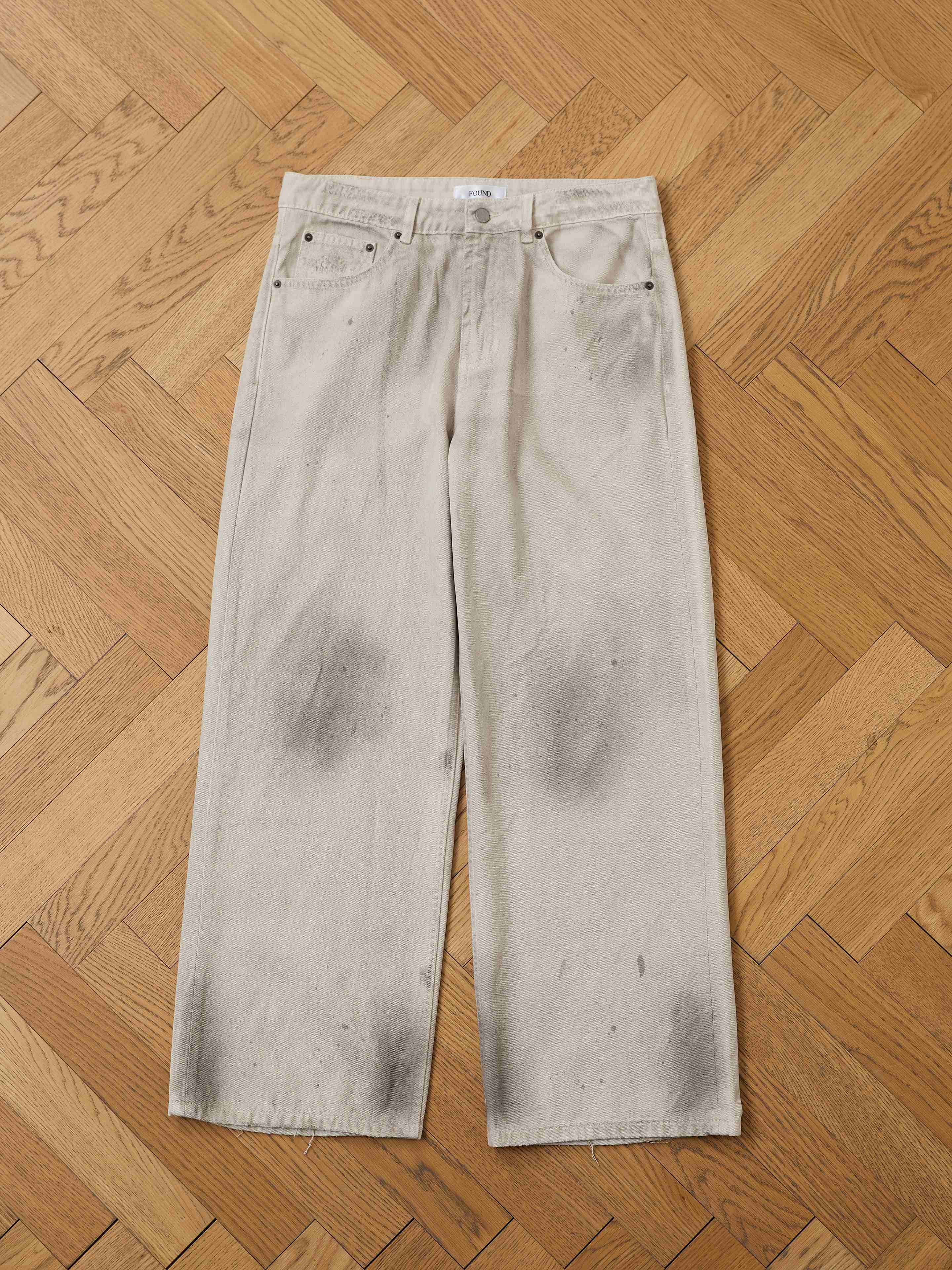 The FOUND Lacy Baggy Jeans feature a relaxed fit and wide-leg design in a distressed concrete wash with visible dark stains, shown laid flat on a wooden parquet floor.