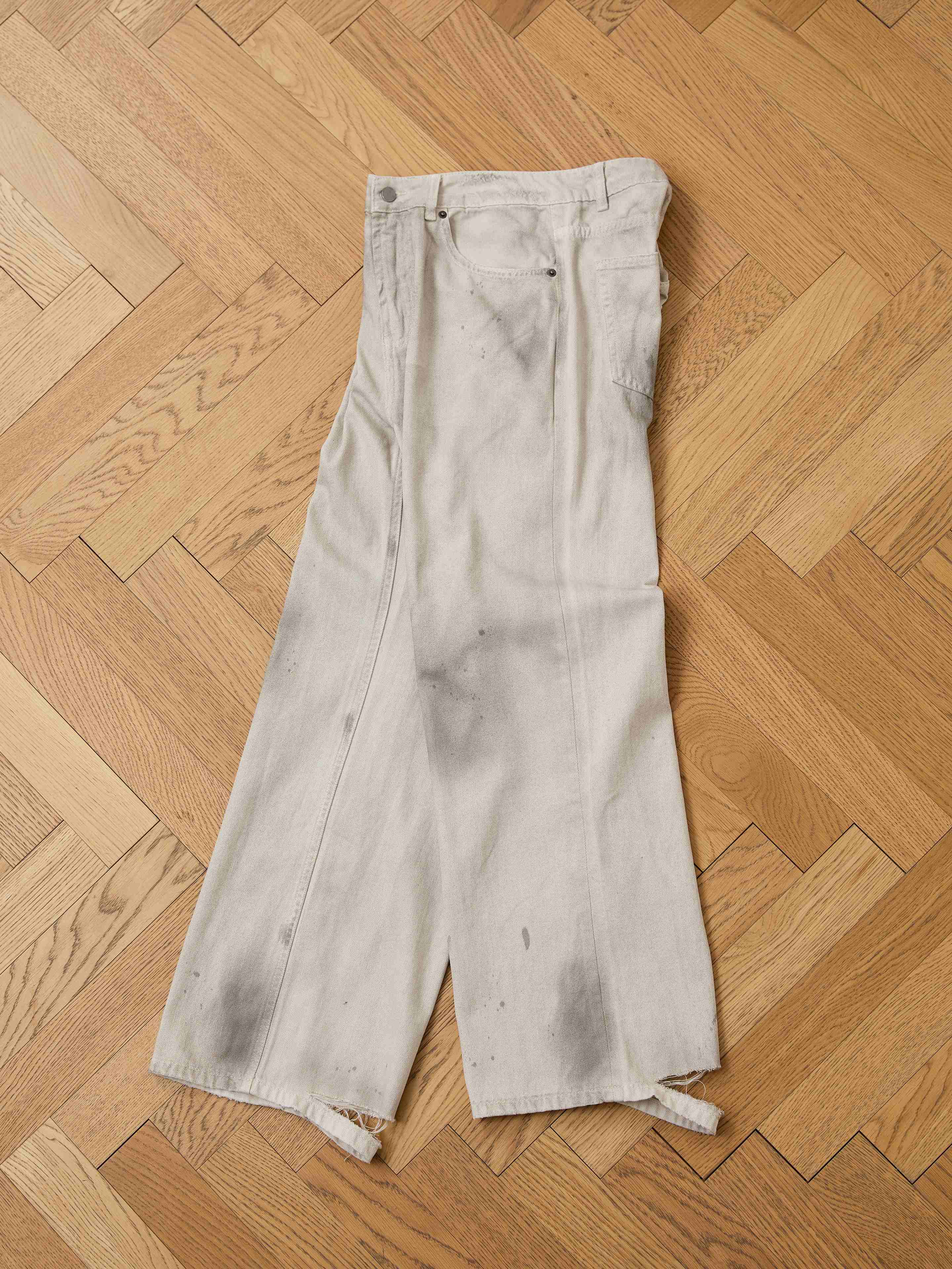 A pair of FOUND Lacy Baggy Jeans featuring a relaxed fit, wide legs, distressed concrete wash, visible stains, and frayed hems lies on a wooden herringbone floor.