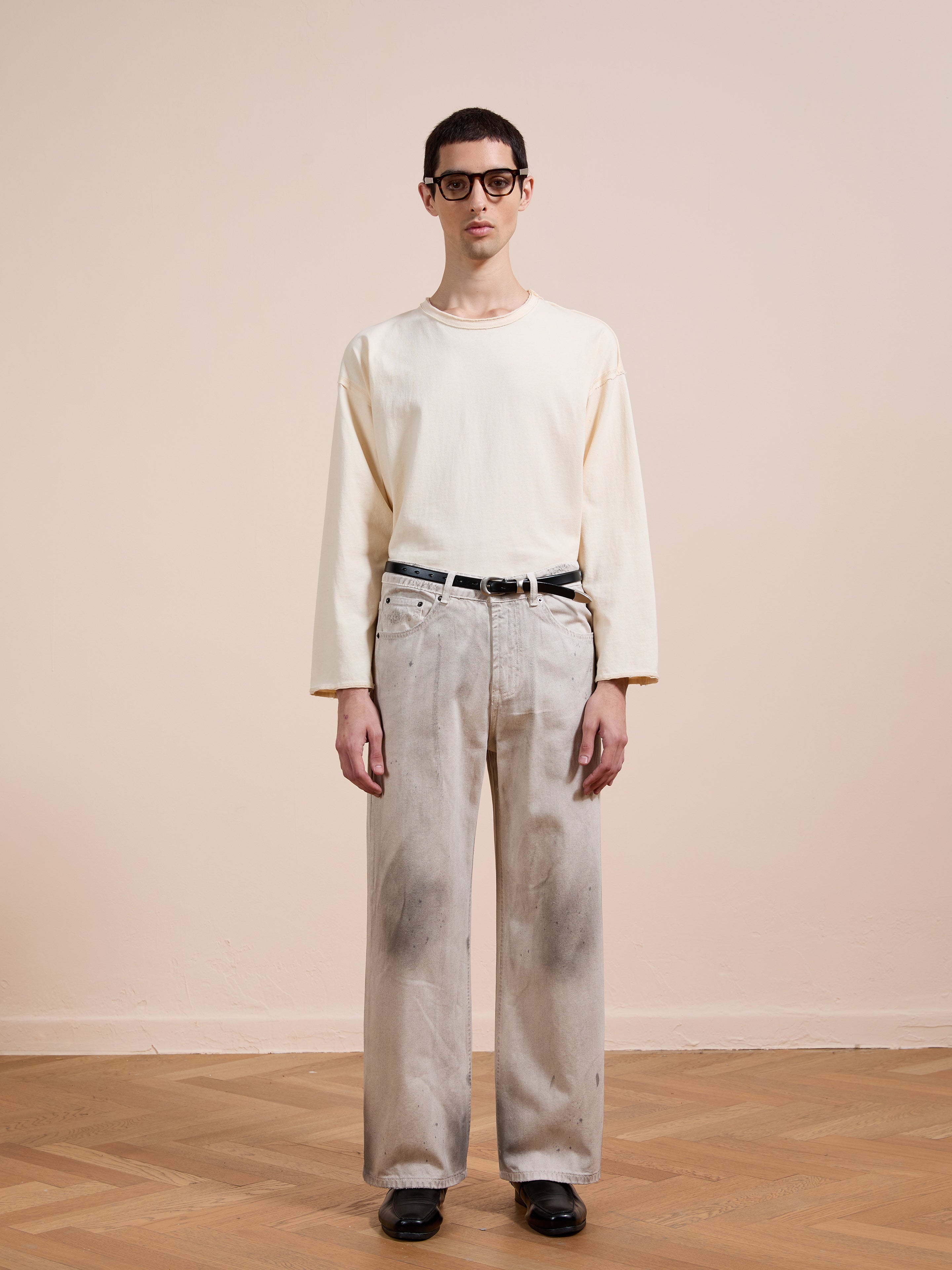 A person stands indoors against a beige wall, wearing glasses, a cream long-sleeve shirt, black belt, black shoes, and FOUND's Lacy Baggy Jeans in a distressed concrete wash.