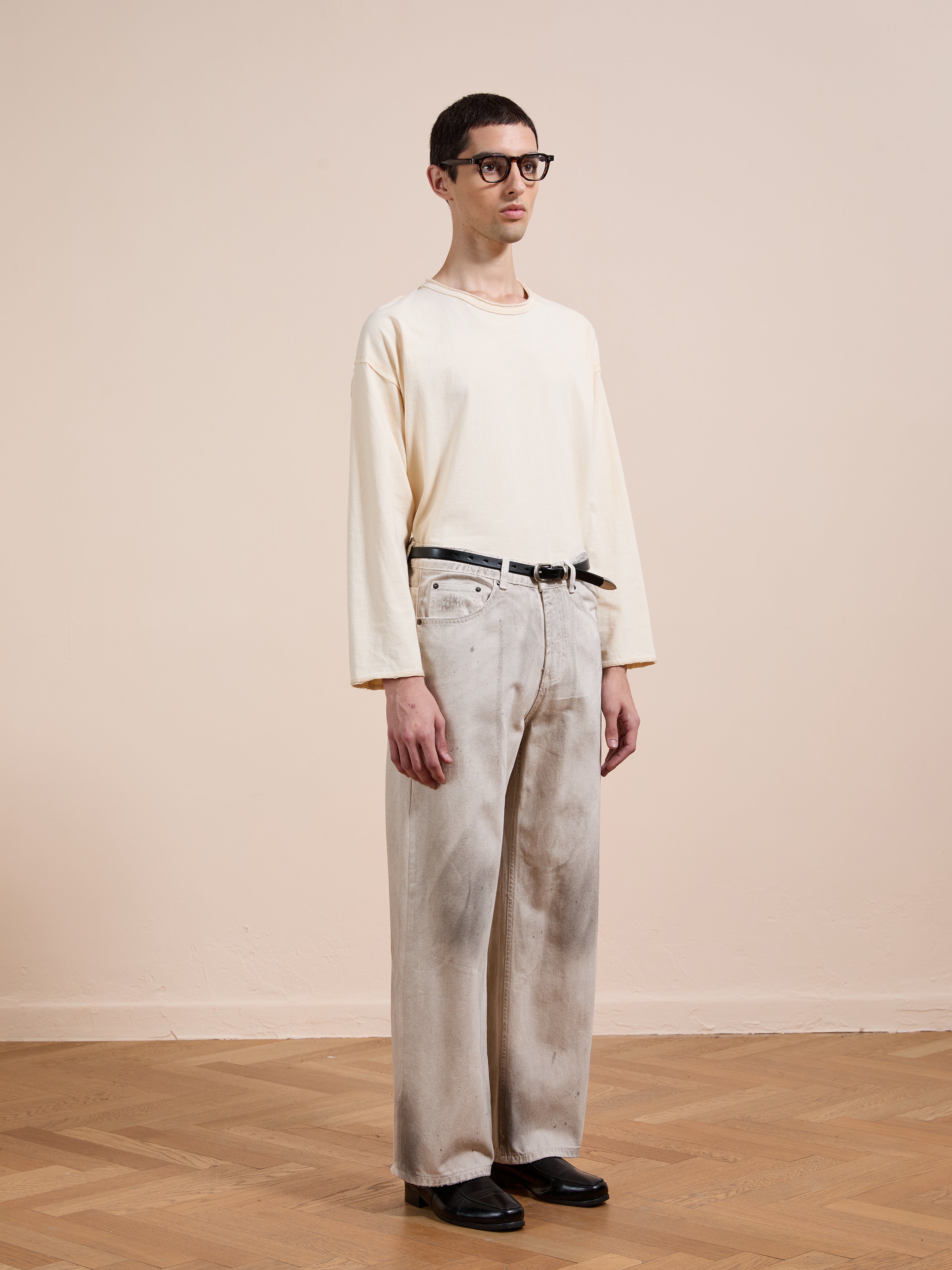 A person with short dark hair and glasses stands indoors in a loose cream long-sleeve shirt, FOUND Lacy Baggy Jeans, a black belt, and black shoes on a wooden floor.