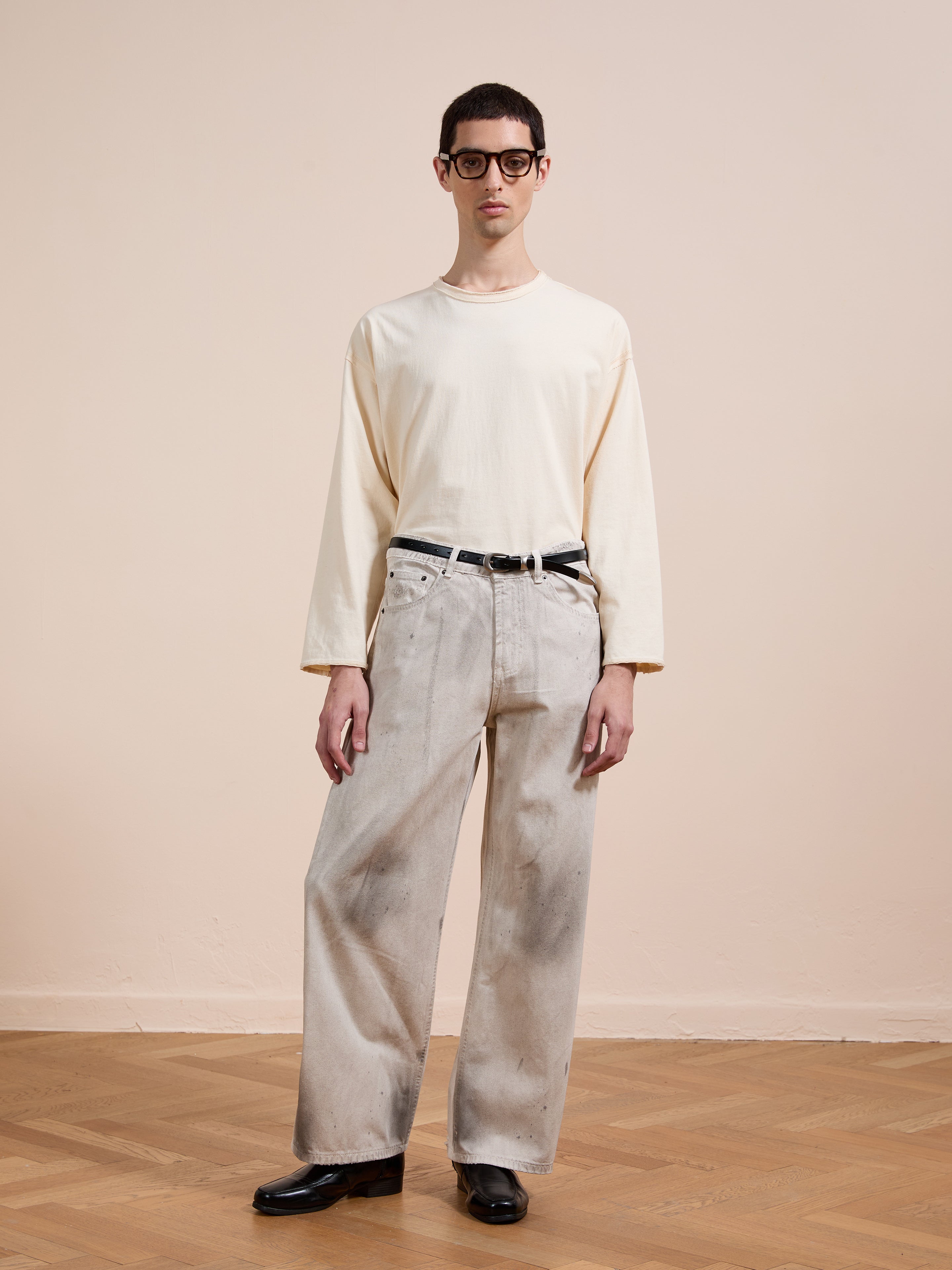 A person in a cream long-sleeve shirt, FOUND Lacy Baggy Jeans in light gray, with a black belt and black shoes stands indoors on a wooden floor against a beige wall.