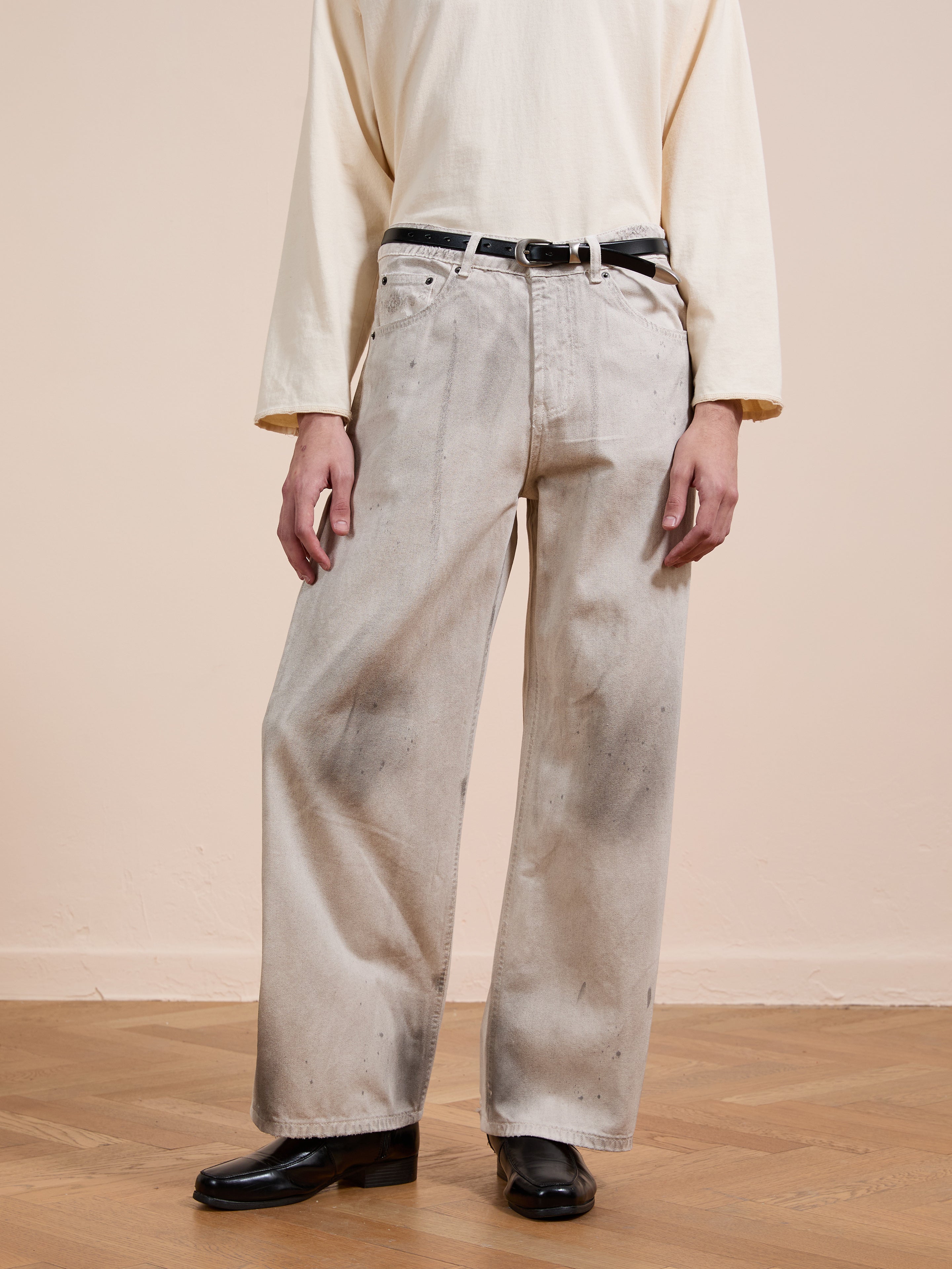 A person wears a loose, long-sleeved cream shirt, FOUND Lacy Baggy Jeans in distressed concrete wash, a black belt, and black shoes, standing on a wooden floor against a pale wall.