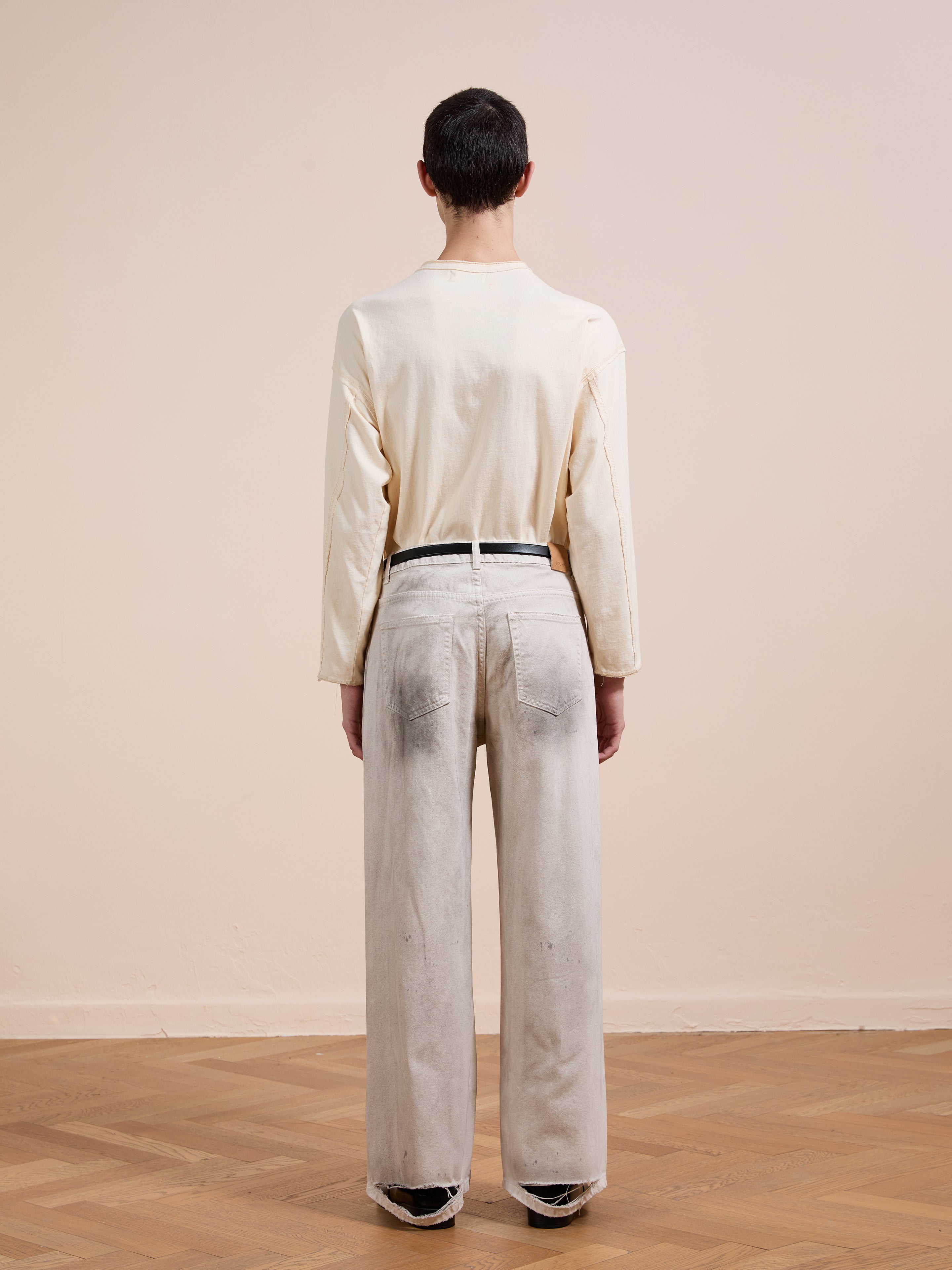 A person stands indoors on a wooden floor, facing away, wearing a cream long-sleeve shirt tucked into FOUND Lacy Baggy Jeans in light gray with a black belt.