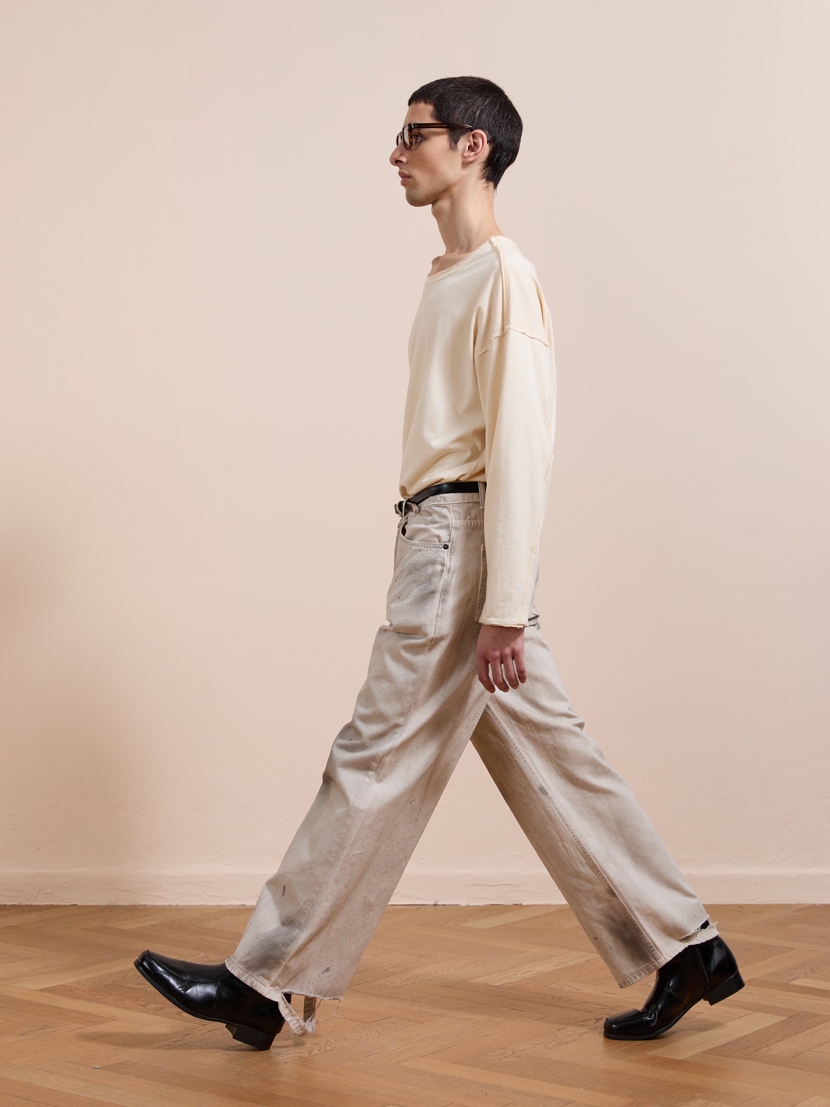 A person with short dark hair, glasses, a cream long-sleeve shirt, black shoes, and FOUND Lacy Baggy Jeans walks indoors on a wooden floor with a beige wall in the background.