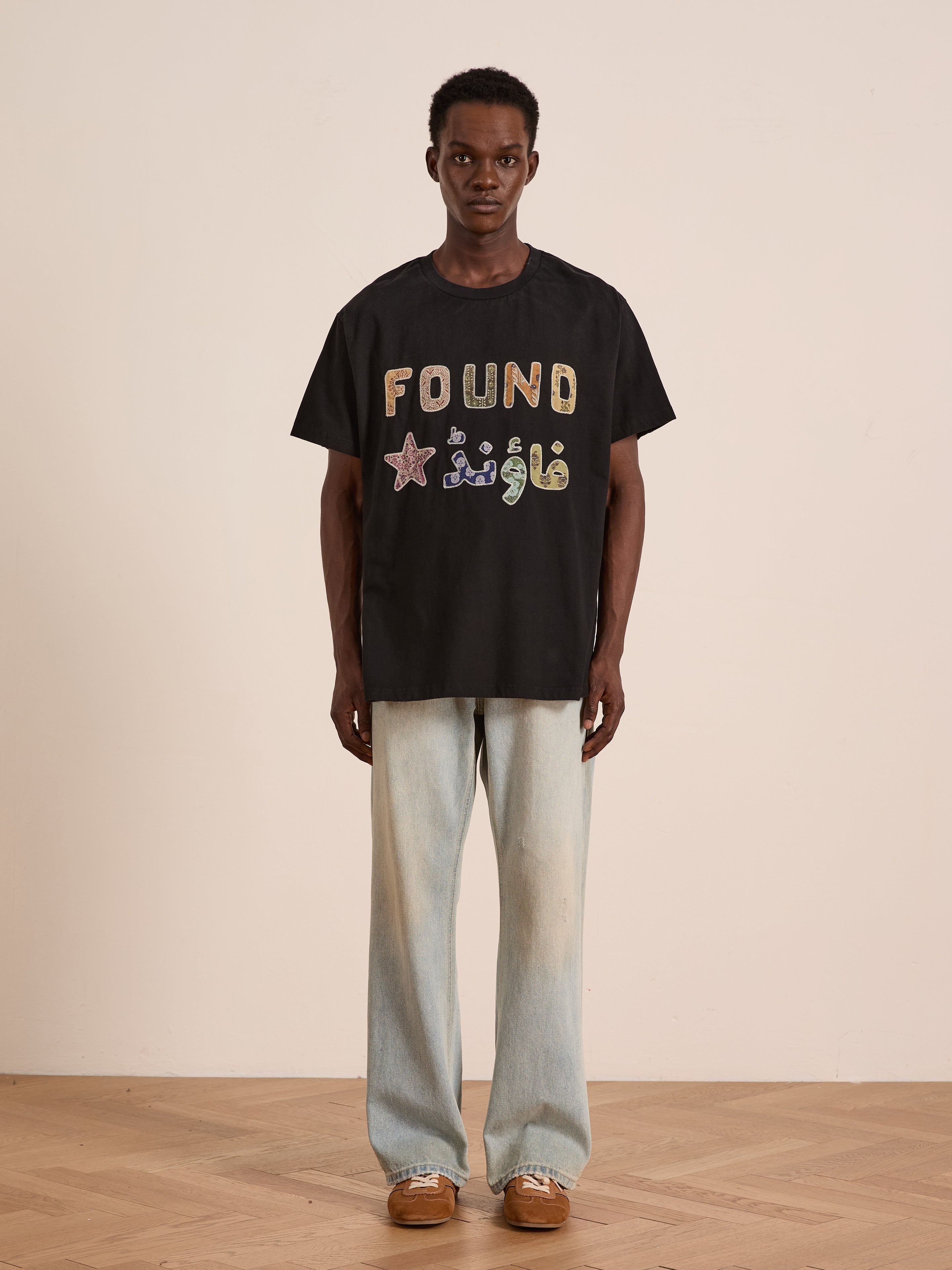 A person stands against a plain wall wearing the FOUND Language Tapestry Logo Tee with colorful symbols, paired with vintage-fade light blue jeans and brown shoes.