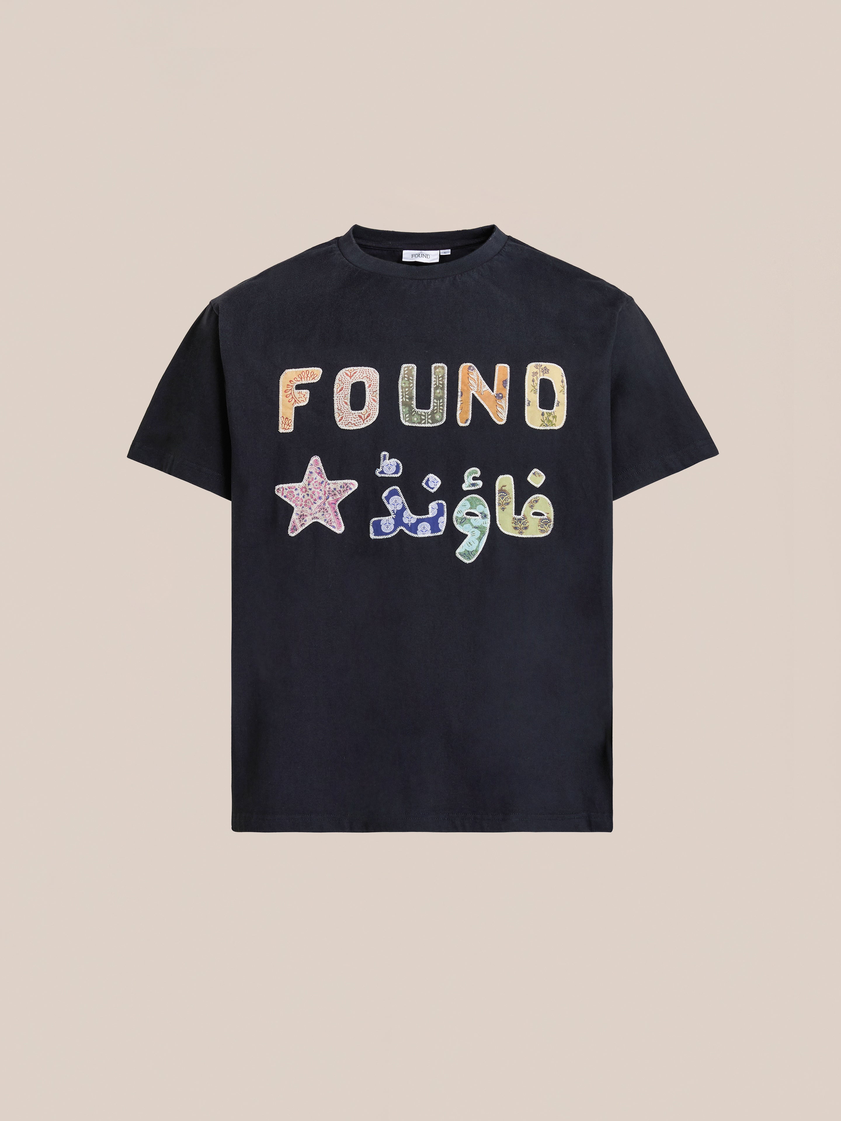 The FOUND Language Tapestry Logo Tee is a black t-shirt with "FOUND" and a star in colorful, vintage-faded lettering (including Arabic script), shown on a plain beige background.