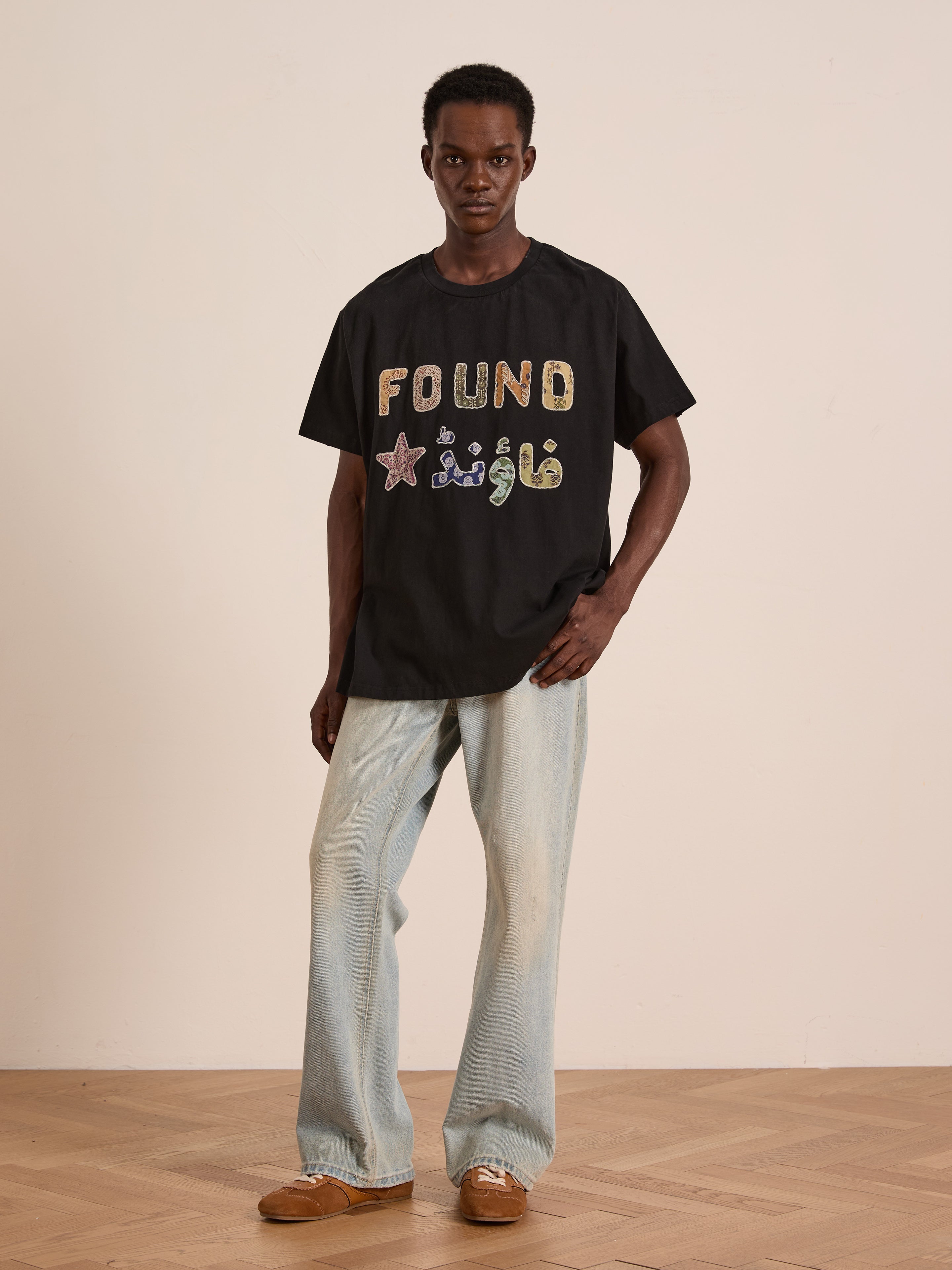 A person stands against a plain wall, wearing the FOUND Language Tapestry Logo Tee in black, light blue vintage-faded jeans, and brown shoes.