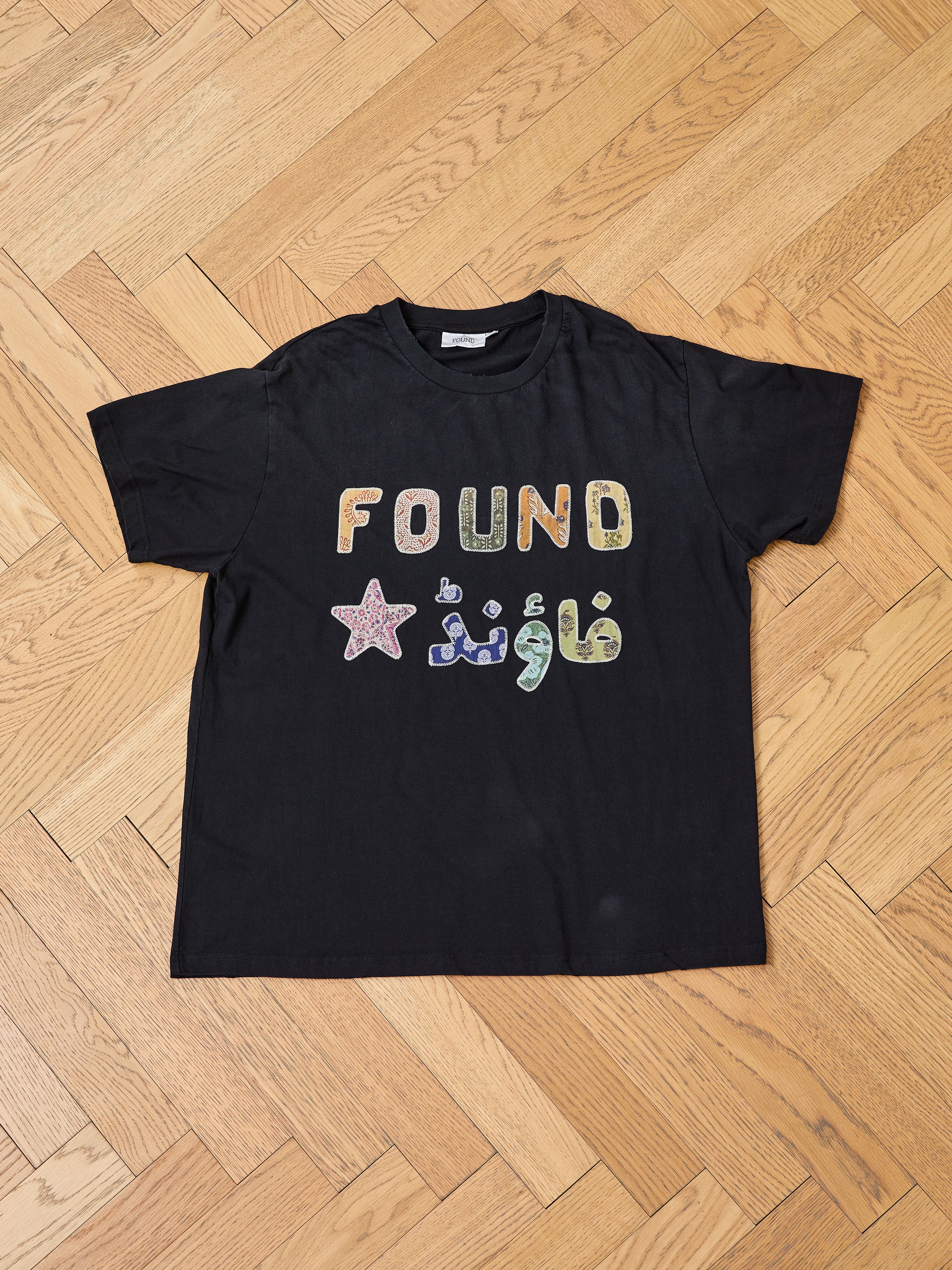 The FOUND Language Tapestry Logo Tee is a black shirt featuring "FOUND," a star, and the Arabic word for "found" in colorful letters with a vintage-inspired fade, shown on a wooden parquet floor.