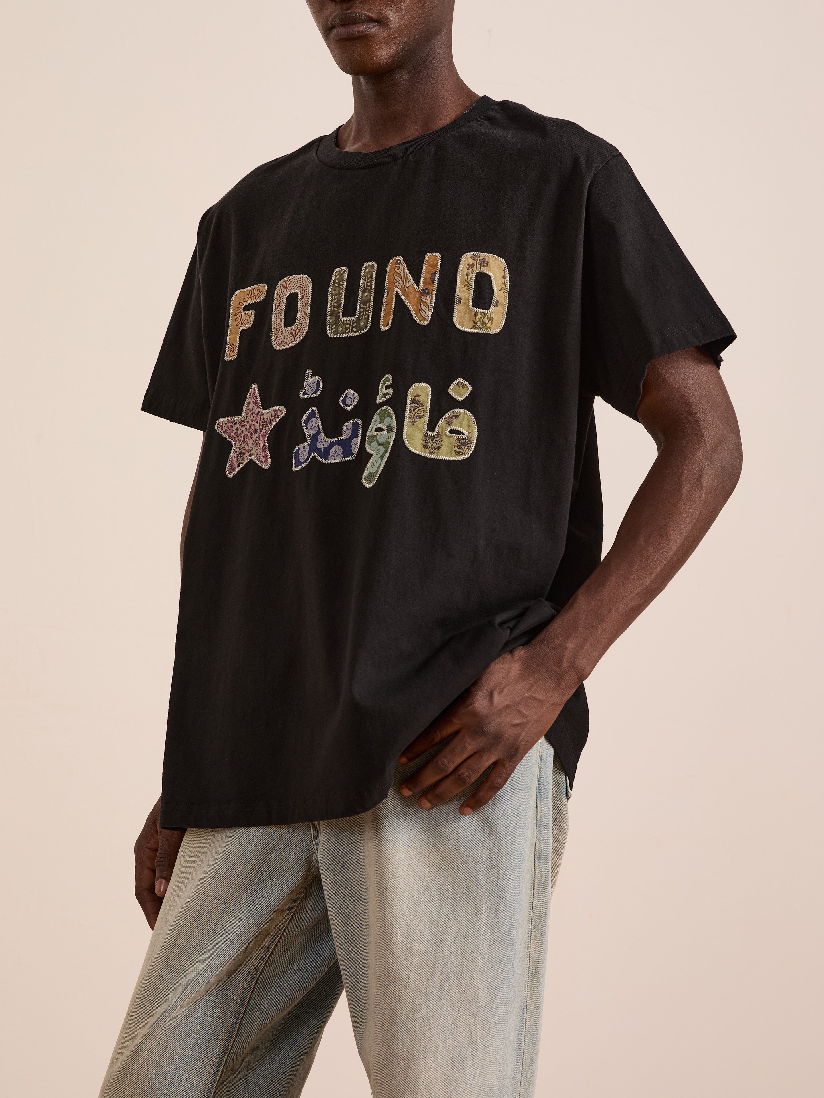 A person wearing the FOUND Language Tapestry Logo Tee—a black shirt with "FOUND" and a star in English and Arabic, vintage-faded—paired with light blue jeans.