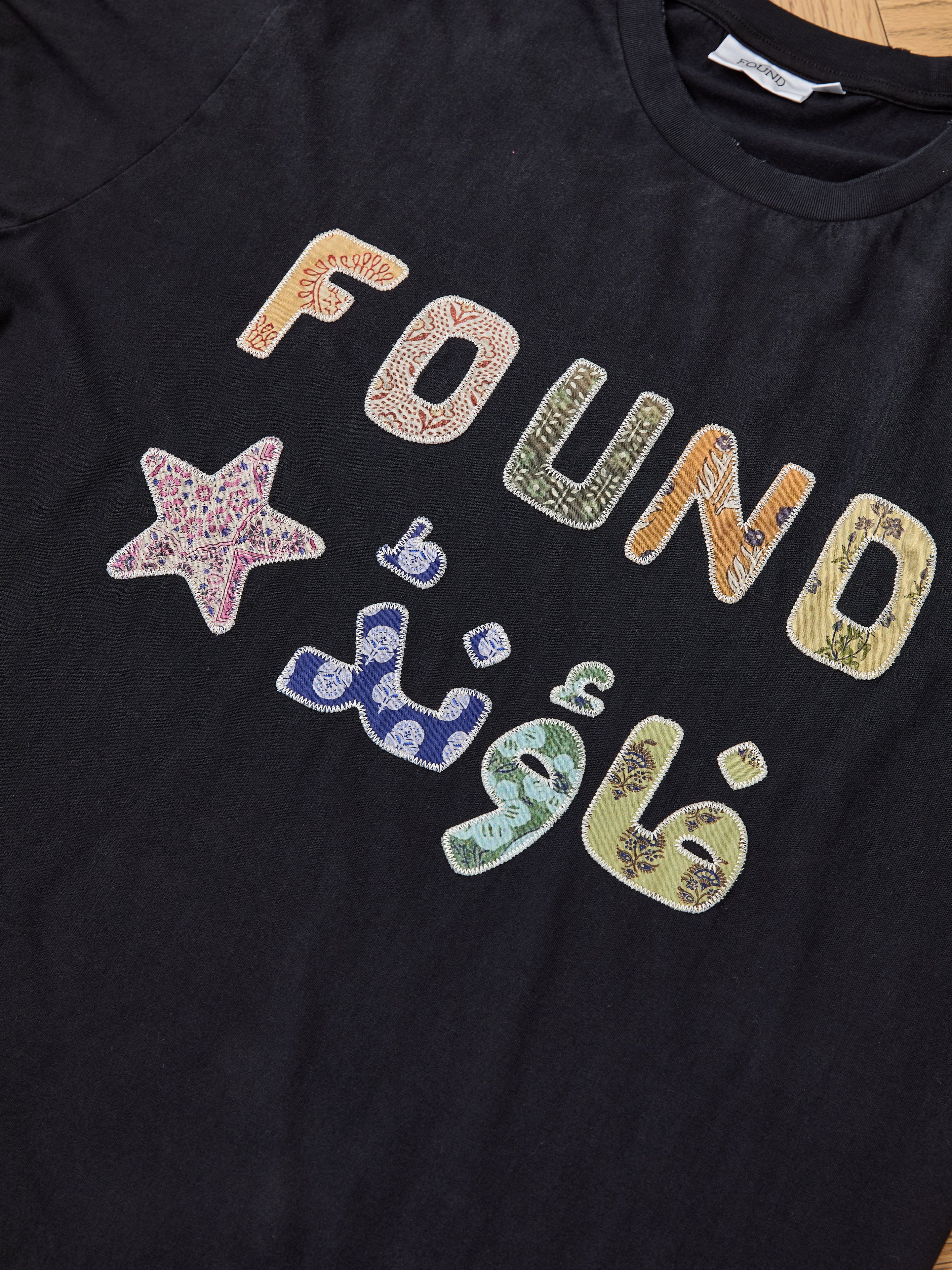 The FOUND Language Tapestry Logo Tee features "FOUND" with a star in colorful, South Asian textiles-inspired patterns, Arabic text below, and vibrant fabric details—all combining for a unique, standout look.
