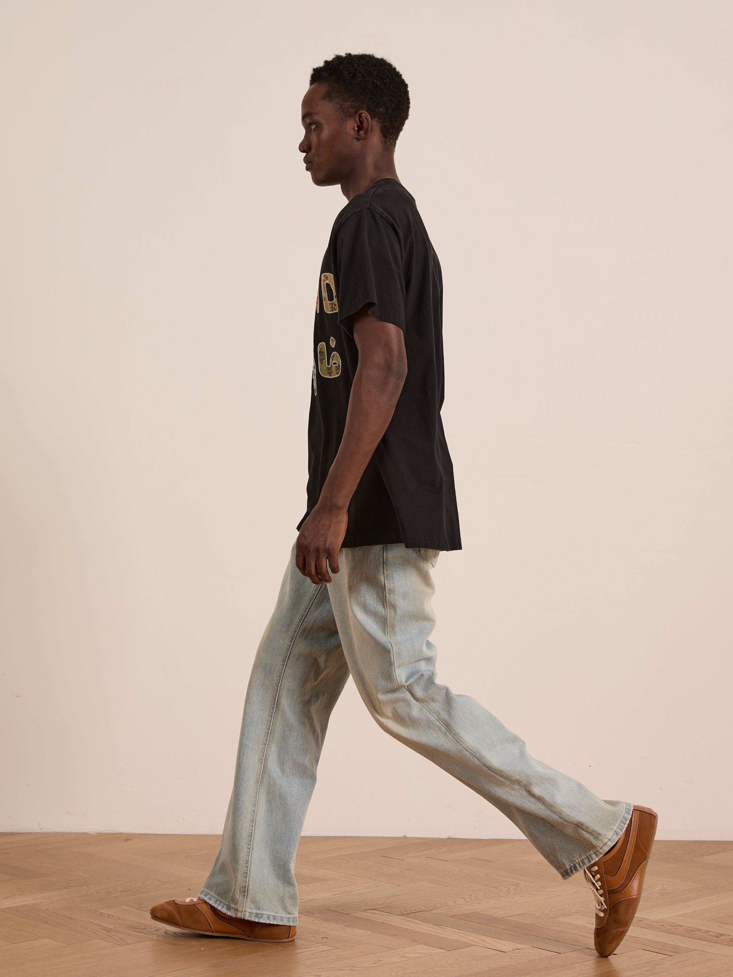 A person wears the FOUND Language Tapestry Logo Tee, light blue jeans, and brown shoes while walking indoors on a wooden floor against a plain light-colored wall.