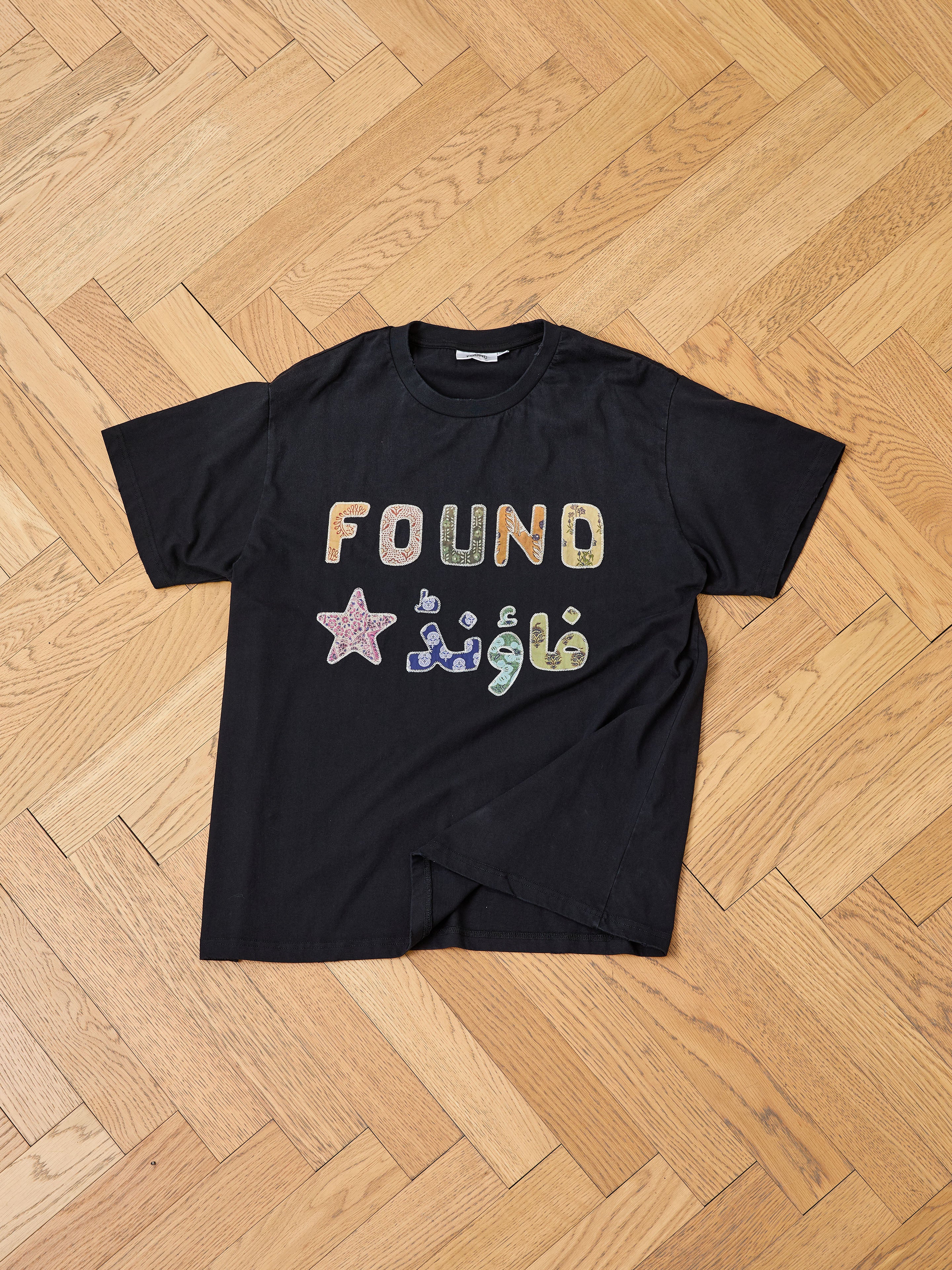 The FOUND Language Tapestry Logo Tee is a black shirt with colorful "FOUND" lettering in English and Arabic, a star accent, and a vintage-inspired fade, shown on a wooden floor.