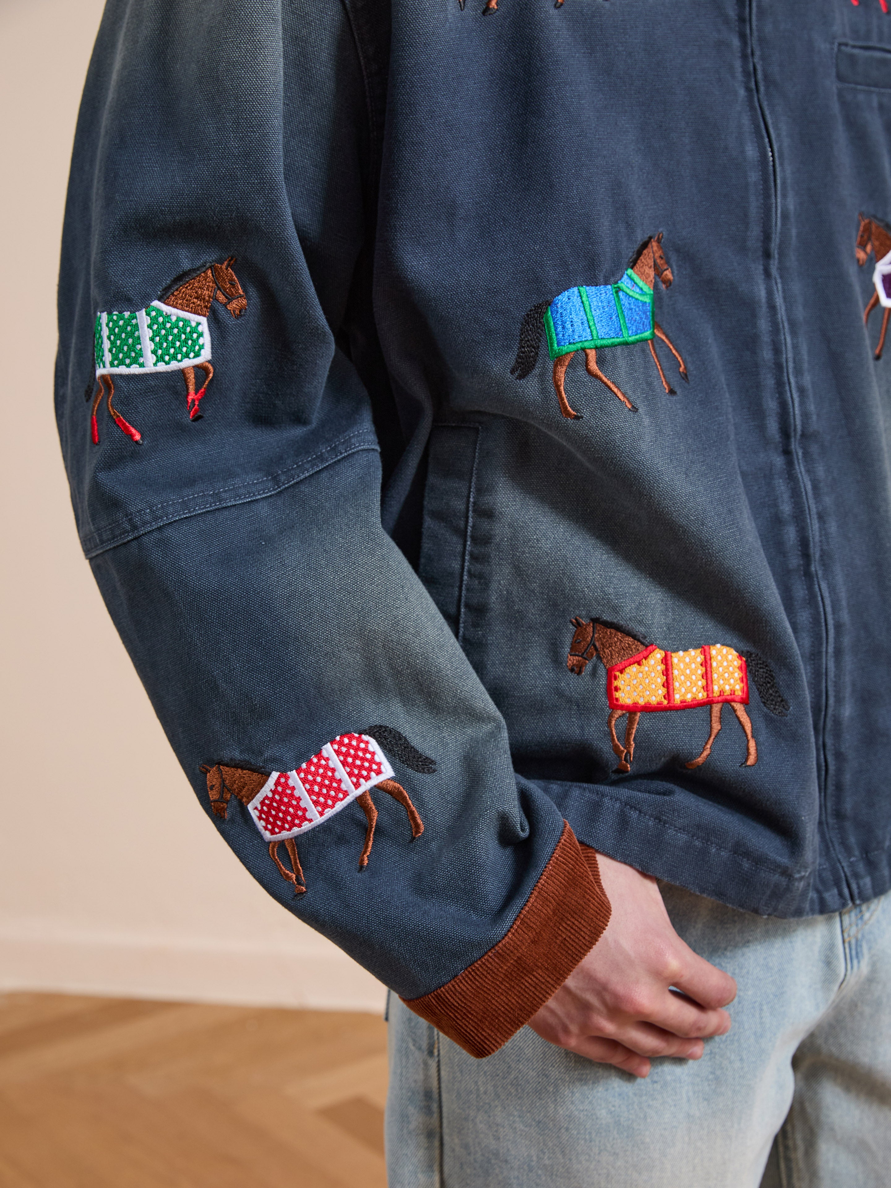 A person wears the Found Lariat Horse Embroidered Work Jacket in unisex navy, featuring vibrant horse embroidery. Their hand rests in their pocket, paired stylishly with light blue jeans.